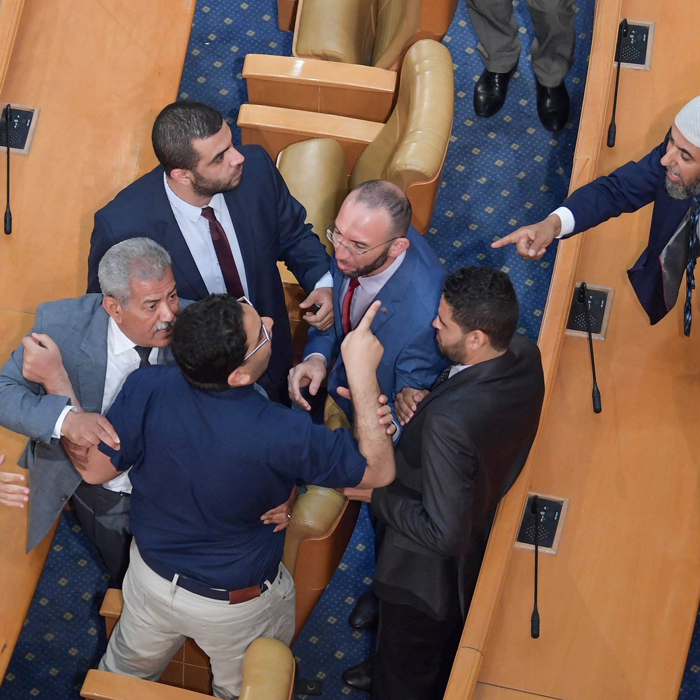 The image shows a heated confrontation occurring in a legislative assembly. Several men are engaged in a tense discussion or argument, with some gesturing animatedly. The setting appears to be a formal chamber, indicated by the rows of seats and the official appearance of the individuals. Some men are standing closely together, while others seem to be pointing or raising their hands, indicating strong emotions. Overall, the scene conveys a moment of conflict or debate among the legislators.