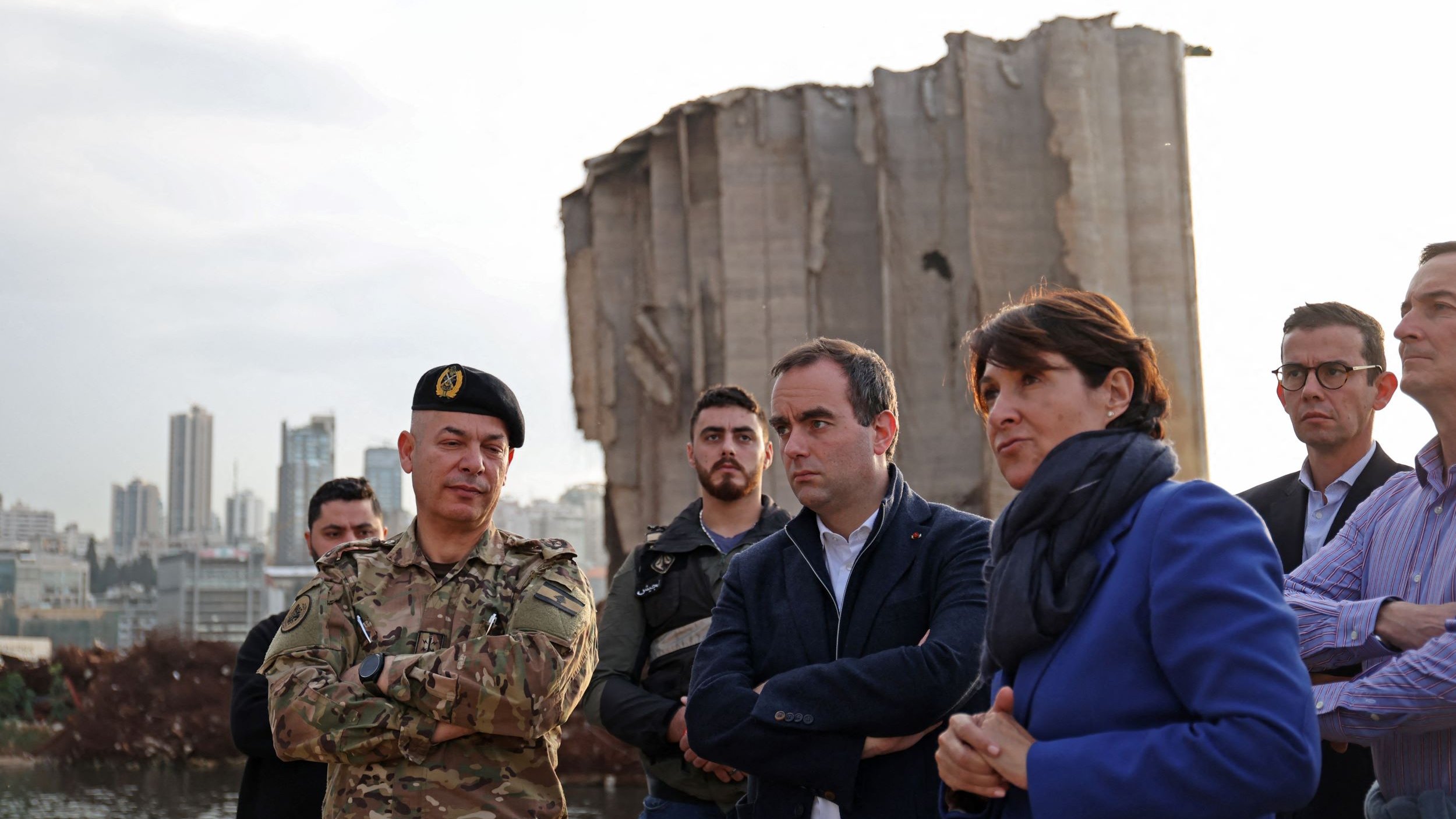 در این تصویر، گروهی از افراد در کنار یک ساختمان ویران، که ظاهراً بخشی از آن تخریب شده، ایستاده‌اند. یک نظامی با لباس نظامی و کلاه فرماندهی در سمت چپ قرار دارد. کنار او مردان و زنانی با لباس‌های رسمی و غیررسمی ایستاده‌اند و به نظر می‌رسد که در حال صحبت و تبادل نظر هستند. پس‌زمینه تصویر نشان‌دهنده شهری با ساختمان‌های بلند است. نور در تصویر کم است و فضای کلی حالت جدی و تاملی دارد.