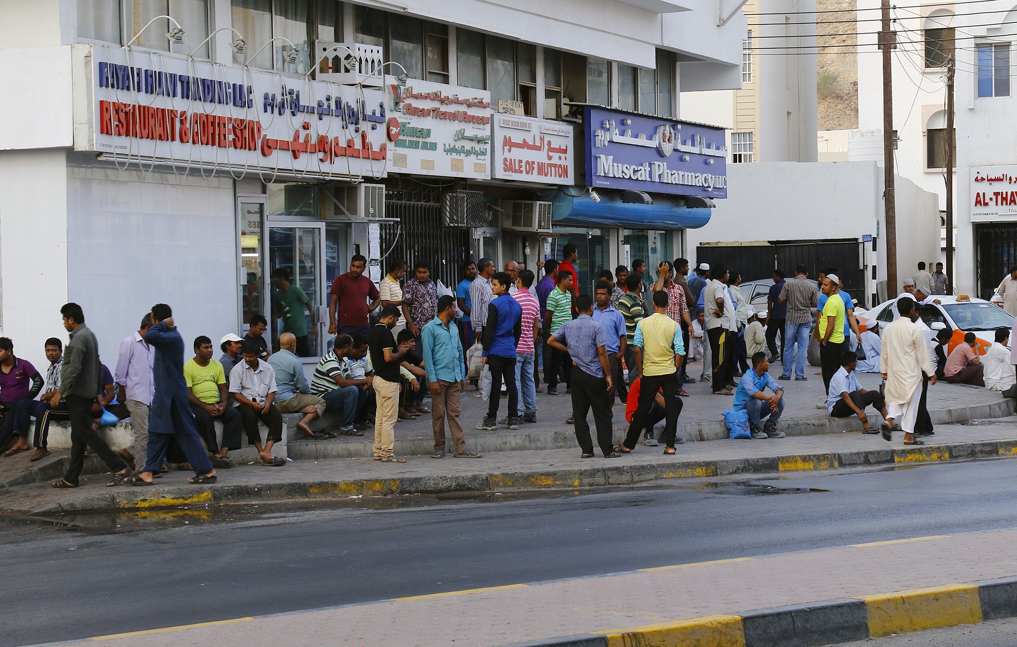 تظهر الصورة مشهداً من شارع في منطقة حضارية مزدحمة. يوجد العديد من الأشخاص يتجمعون بالقرب من storefrontات ومتاجر متنوعة. يمكن رؤية مجموعة من الرجال والمارة يجلسون أو يقفون، وبعض الناس مشغولون بالتحدث مع بعضهم. يضم المشهد أيضاً لافتات باللغة العربية تشير إلى المتاجر والمرافق. الجو يبدو نشطًا، مع حركة مرور على الشارع في الخلفية.