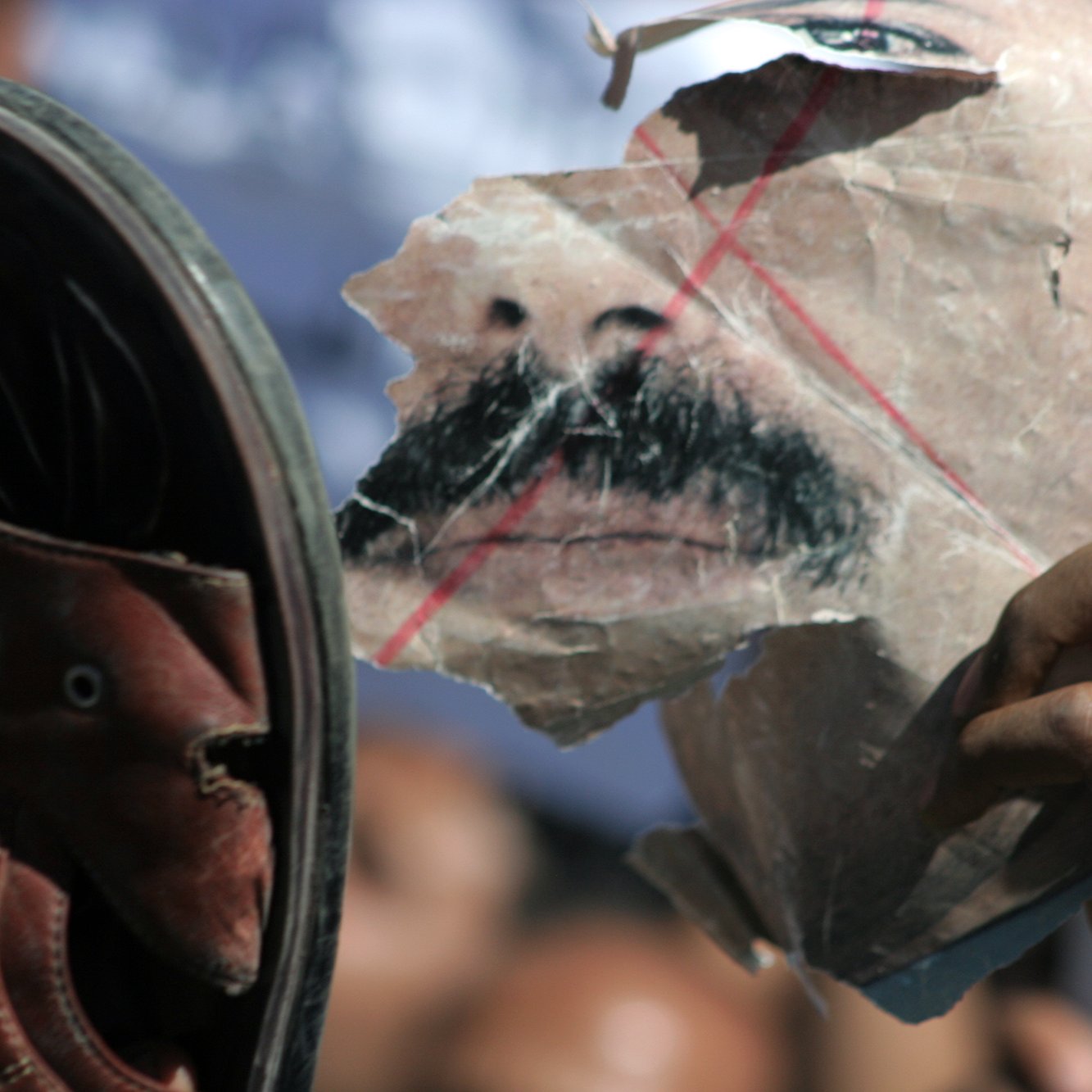 L'image montre une chaussure levée vers un visage déchiré, probablement représentant une personne. Le visage est marqué par des lignes rouges et semble avoir été martelé ou abîmé. L'atmosphère de l'image suggère de la contestation ou de l'opposition, avec une foule floue en arrière-plan. La chaussure et le visage déchiré symbolisent peut-être un acte de rébellion ou de rejet.