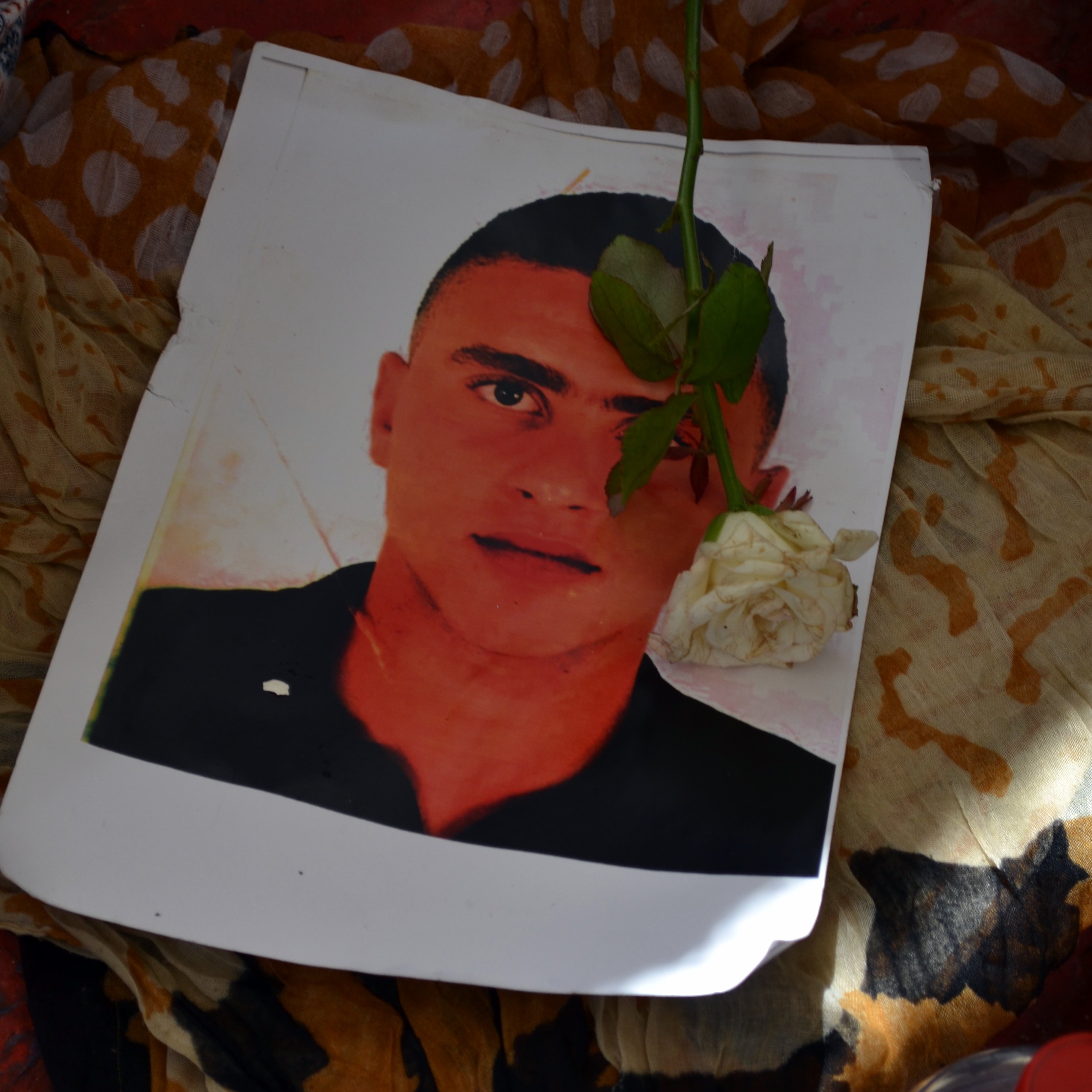 L'immagine mostra un ritratto di un giovane uomo posato su un tessuto colorato. Sopra il ritratto è adagiata una rosa bianca, che aggiunge un tocco di dolcezza e commemorazione. Il contrasto tra il volto del ragazzo e il fondo morbido e multicolore crea un'atmosfera di riflessione e rispetto.