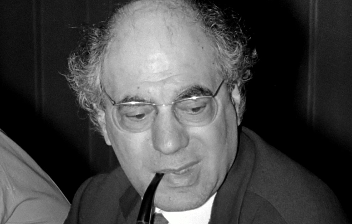 تظهر في الصورة رجل يجلس على طاولة، يبدو أنه في وضعية نقاش أو اجتماع. لديه شعر رمادي ومجعد، ويرتدي نظارات ويشعل غليونًا. يرتدي بدلة داكنة مع قميص وروابط. تعكس تعابير وجهه تركزًا واهتمامًا. البيئة المحيطة تبدو بسيطة، مع تفاصيل غير واضحة في الخلفية.