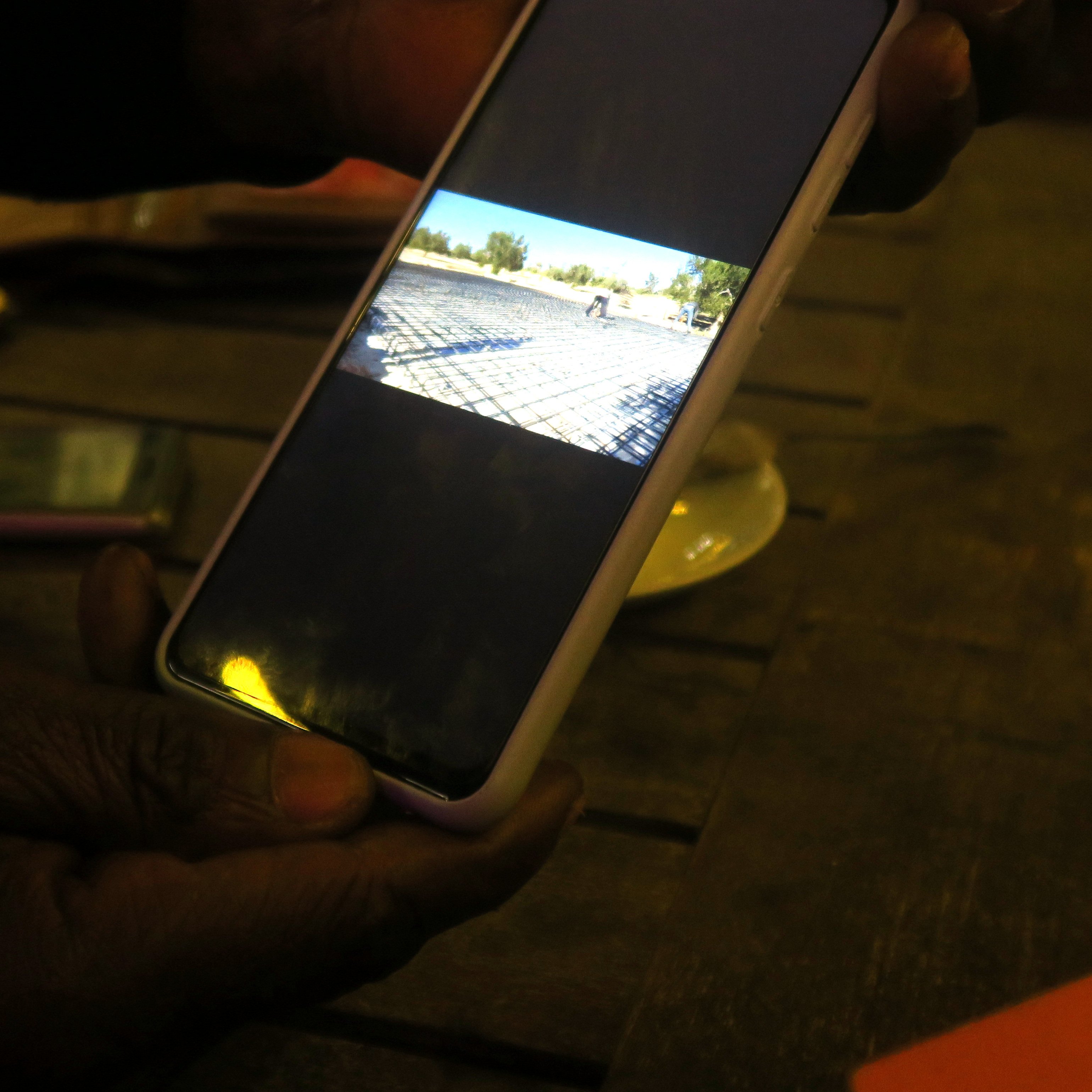 L'image montre une main tenant un téléphone mobile qui affiche une photo sur son écran. La photo visible montre un paysage avec un chemin de pavés et un véhicule au loin. L'éclairage est faible, ce qui donne une ambiance douce à la scène. Le téléphone est positionné sur une table en bois, et l'arrière-plan contient des objets flous, suggérant un environnement convivial.