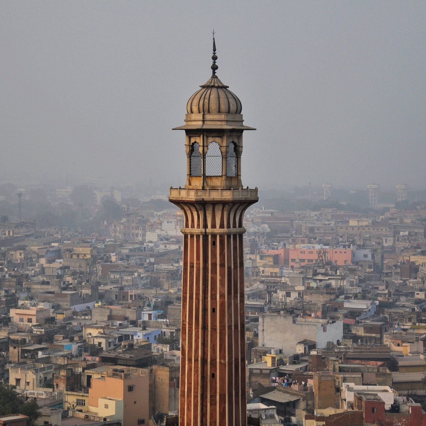 L'image montre un paysage urbain avec une tour élancée qui se dresse au milieu de nombreux bâtiments. La tour a une architecture distinctive, avec des motifs ornés et un dôme en haut. En arrière-plan, on peut voir une vaste étendue de maisons et de structures, témoignant d'une villeDense. L'atmosphère semble légèrement brumeuse, suggérant une pollution ou une humidité ambiante. Les couleurs des bâtiments varient, ajoutant une touche de diversité à l'ensemble de la scène.