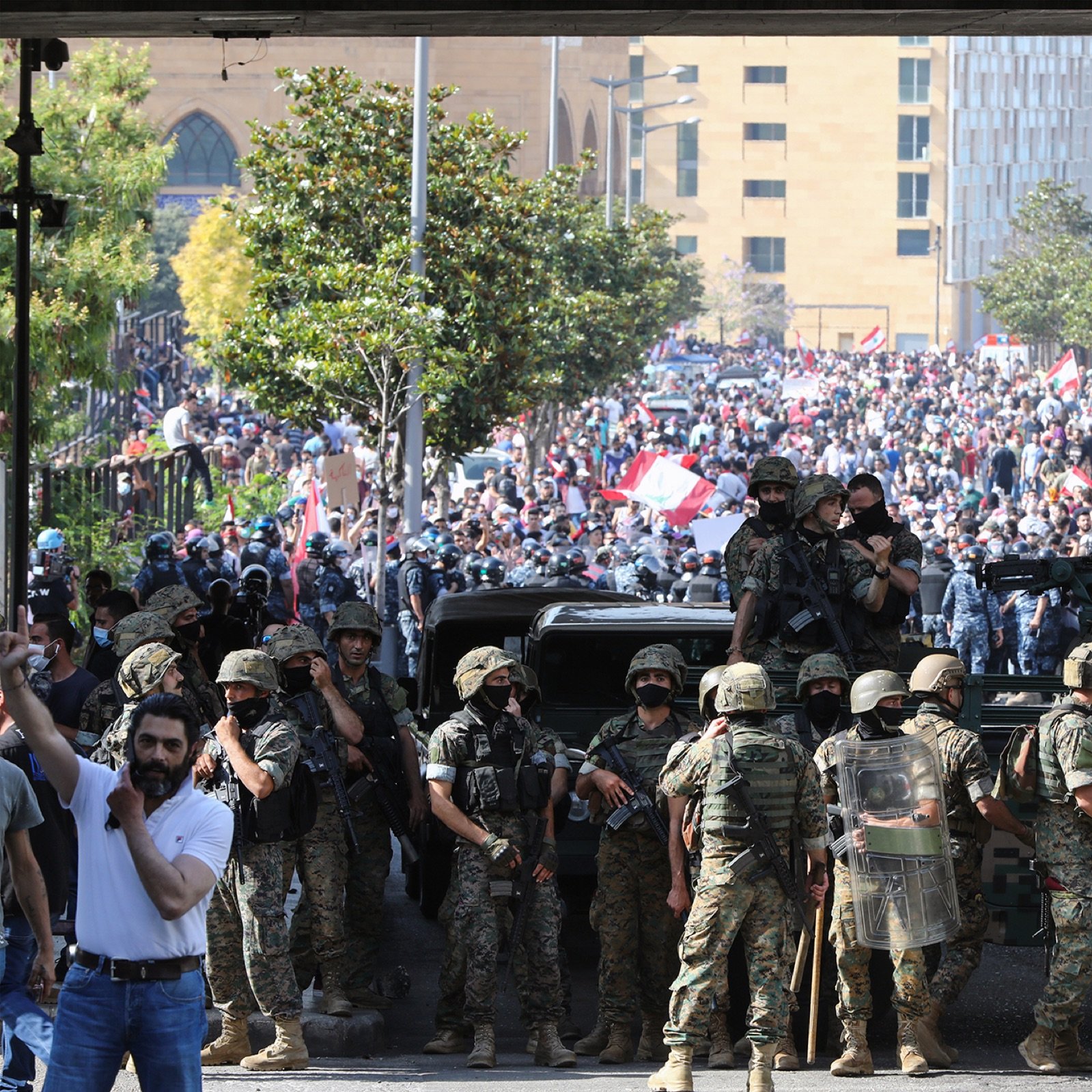 L'image montre une scène de protestation dans une ville, probablement à Beyrouth. Au premier plan, des soldats en uniforme sont alignés, portant des équipements de protection et des casques. Ils semblent faire face à une grande foule de manifestants qui se trouvent derrière eux. La foule est chargée d'énergie et porte des drapeaux, ce qui indique qu'il s'agit d'une manifestation pour une cause. Au fond, on peut voir des bâtiments urbains, ce qui ajoute un contexte à la scène. L'atmosphère est tendue, reflétant un moment significatif dans une dynamique sociale ou politique.