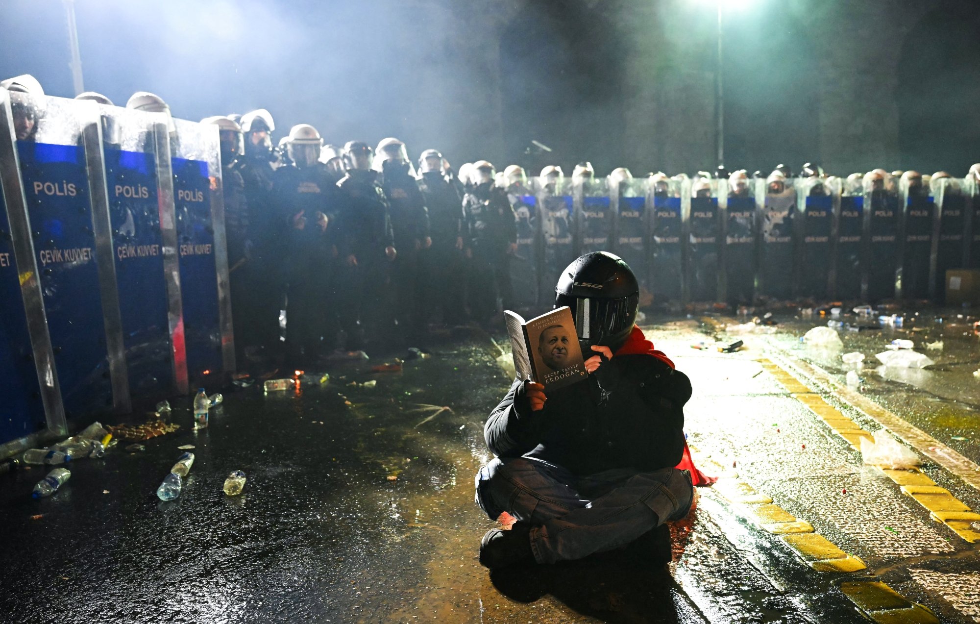 Un manifestant, masqué, lit un livre, face à des policiers en tenue anti-émeute.