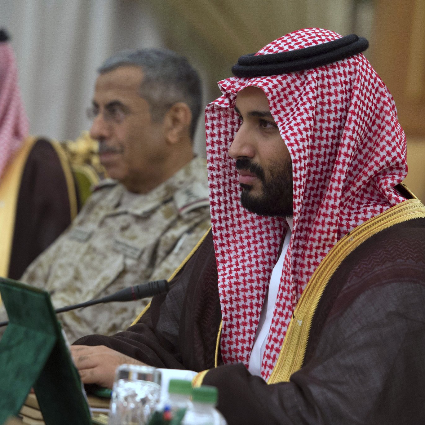 L'image montre un groupe de personnes en tenue traditionnelle. Au premier plan, un homme est assis, portant une agal et un keffieh, souvent associés à la culture saoudienne. Il concentre son attention sur quelque chose devant lui. En arrière-plan, d'autres hommes sont présents, dont l'un porte un uniforme militaire. Les participants semblent être réunis pour une discussion formelle, peut-être lors d'une conférence ou d'un sommet. Des drapeaux et des documents sont visibles sur la table. L'environnement dégage une atmosphère de sérieux et de formalité.