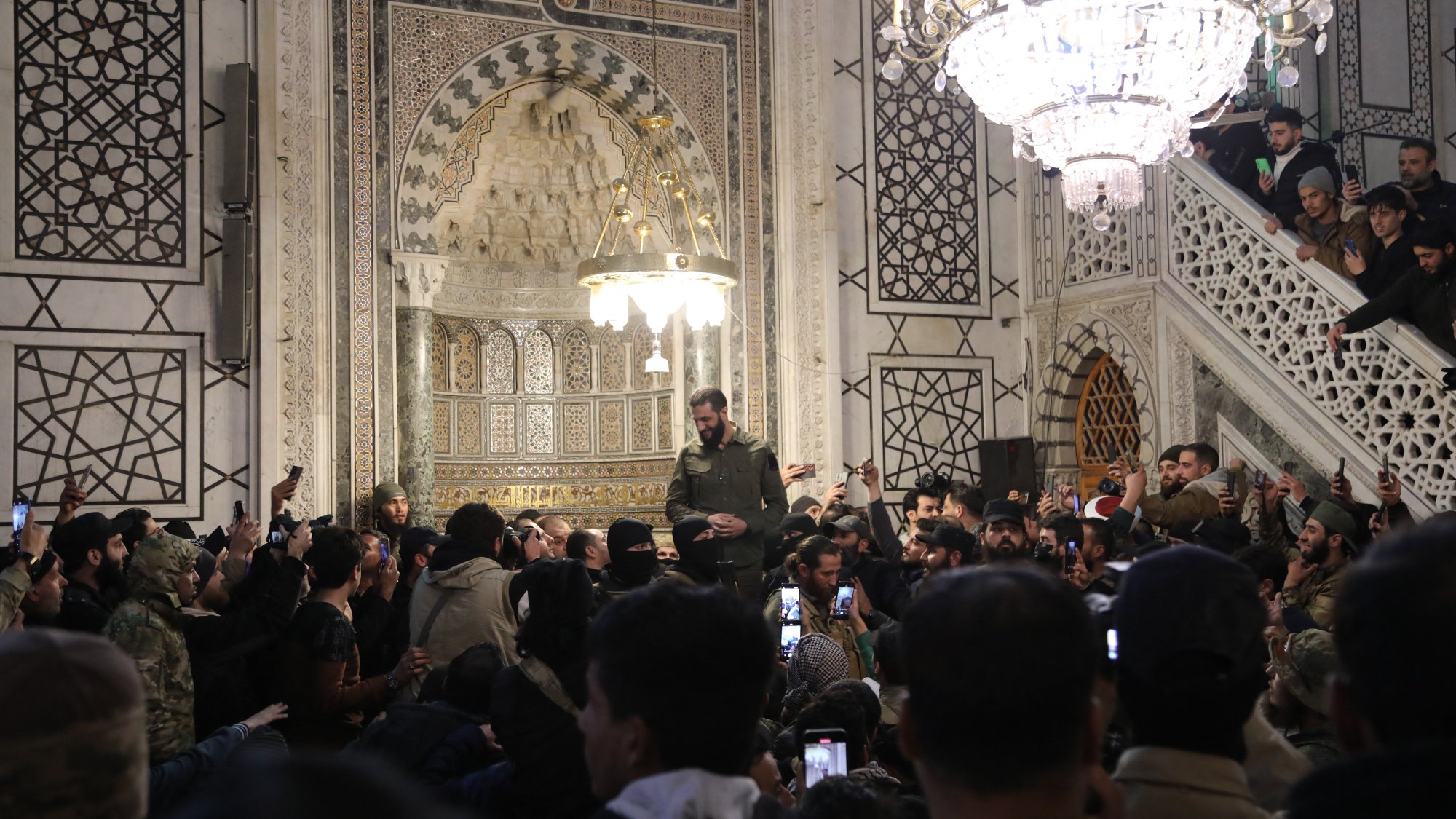 L'image montre un intérieur richement décoré, probablement d'un bâtiment historique ou religieux, avec des motifs géométriques et des ornements élaborés. Au centre, une personne se tient en hauteur, entourée d'une foule qui utilise des téléphones pour capturer le moment. Des lumières brillantes, notamment un lustre, illuminent la scène. L'atmosphère semble être celle d'un rassemblement ou d'un événement significatif.