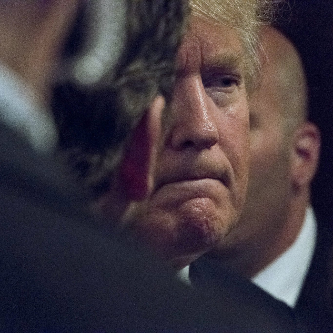 L'image montre un homme avec des cheveux blonds, affichant une expression pensive ou sérieuse. Il est partiellement obscurci par des personnes en arrière-plan, ce qui crée une atmosphère de concentration ou d'intensité. On peut également remarquer des costumes sombres, suggérant un cadre formel.
