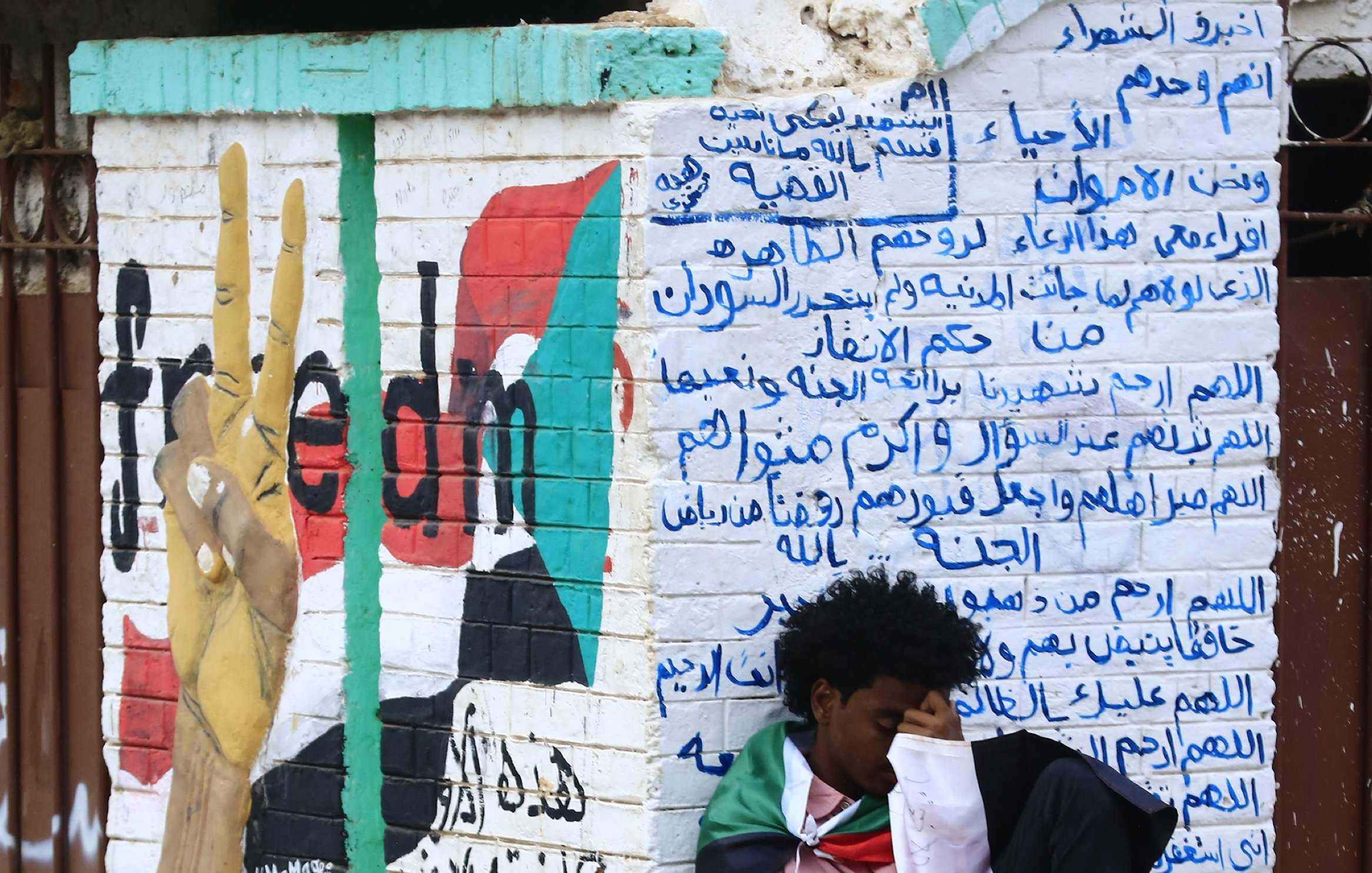 L'image montre un mur peint avec un symbole de paix, probablement représentant un mouvement pour la liberté. À gauche, il y a un graffiti d'une main levant deux doigts, symbolisant le mouvement pacifiste. Sur le mur, des inscriptions en arabe sont visibles, évoquant probablement des messages politiques ou sociaux. Au premier plan, un jeune homme est assis, enroulé dans un drapeau, semblant pensif ou mélancolique. L'atmosphère suggère un moment de réflexion sur des luttes pour la liberté.