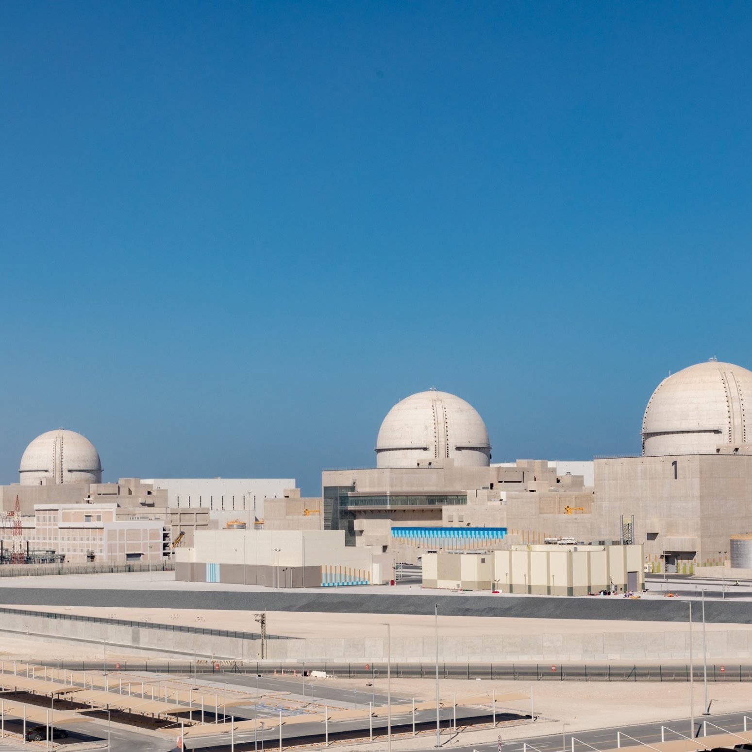 الصورة تظهر منشأة نووية كبيرة، تتكون من عدد من المباني، بعضها يحتوي على قباب كبيرة. السماء في الصورة تبدو زرقاء وصافية، مما يعكس بيئة نظيفة. تصميم المنشأة يبدو حديثًا وعصريًا، مع استخدام الألوان الفاتحة في البناء. المنطقة المحيطة بالمنشأة تبدو مرتبة ومنظمة، مع وجود مساحات واسعة.