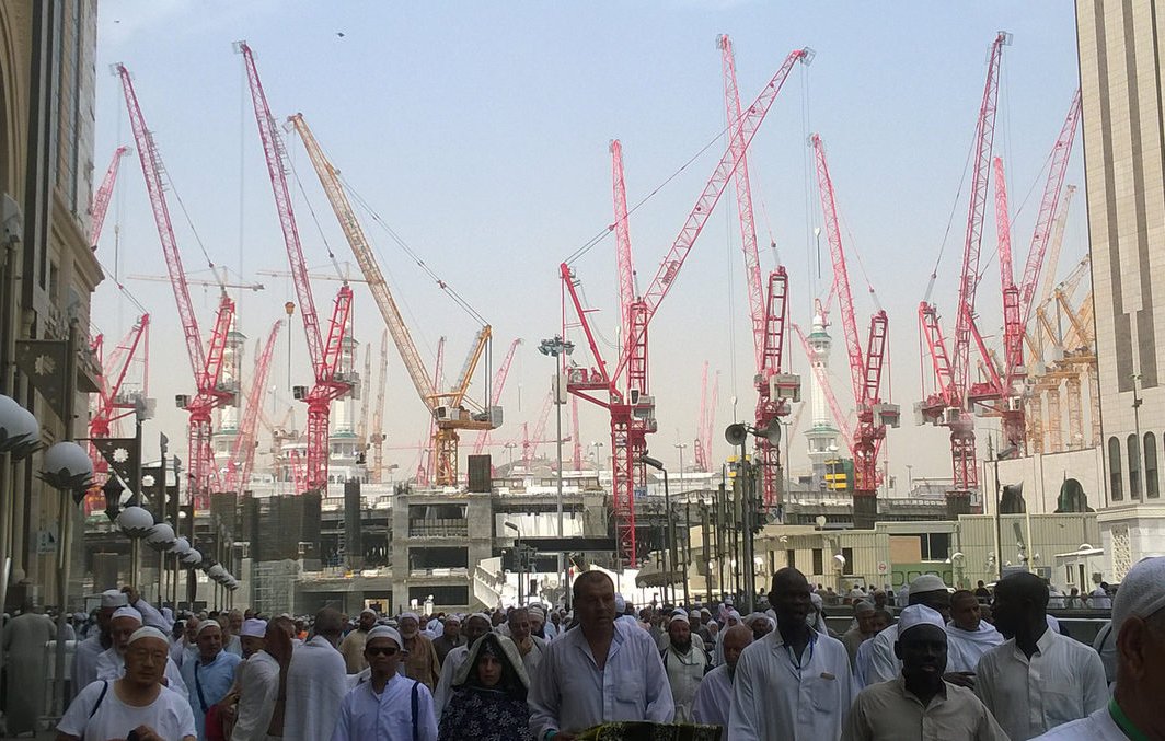 L'image montre un grand nombre de personnes, probablement des pèlerins, se déplaçant dans une zone de construction. En arrière-plan, on peut voir de nombreuses grues rouges qui soulignent les travaux en cours. Les pèlerins portent des vêtements traditionnels, souvent blancs, ce qui est courant lors des pèlerinages. L'environnement semble animé, avec une ambiance de mouvement et d'activité en raison des constructions.