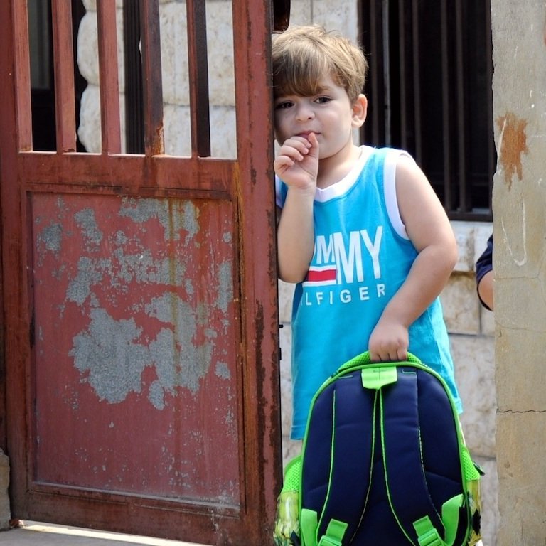L'image montre un jeune garçon se tenant dans l'encadrement d'une porte. Il porte un débardeur bleu avec des inscriptions et tient un sac à dos coloré. Le garçon semble pensif, en train de sucer son pouce. La porte derrière lui est rustique et en métal, avec des jeux d'ombre et de lumière. L'environnement semble simple et authentique, suggérant une ambiance tranquille.