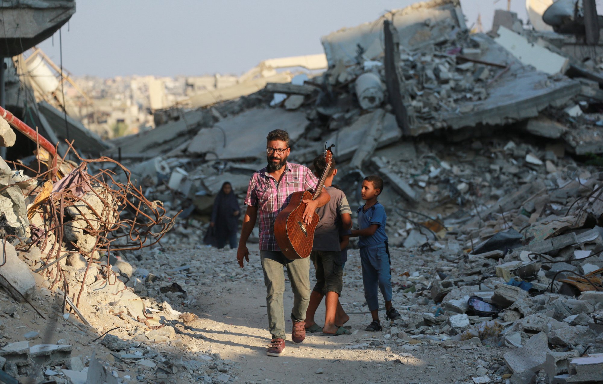 L'image montre un homme marchant à travers des décombres, tenant une guitare. Il est accompagné d'un jeune garçon. L'environnement est en ruines, avec des débris et des structures effondrées autour d'eux. L'atmosphère semble à la fois désolée et résiliente, illustrant un contraste entre la musique et la destruction.