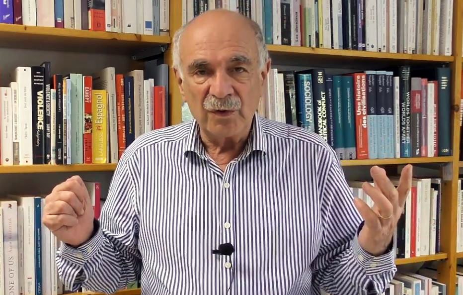 L'image montre un homme debout devant une étagère remplie de livres. Il porte une chemise à rayures et semble s'adresser à un public ou donner une présentation. Ses mains sont levées, ce qui indique qu'il est en train de faire un point ou d'expliquer quelque chose. L'arrière-plan est constitué de livres aux couvertures variées, suggérant un environnement intellectuel ou académique.