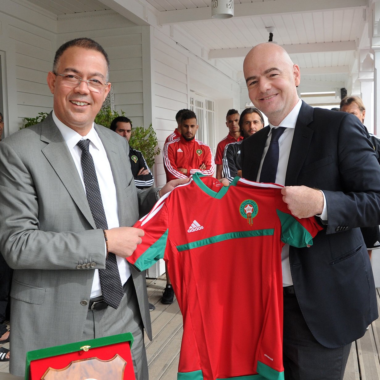 L'image montre deux hommes souriants qui se tiennent devant un groupe de personnes en arrière-plan. L'un d'eux porte un costume gris et tient un maillot de football rouge et vert, probablement aux couleurs de l'équipe nationale marocaine. L'autre homme, en costume sombre, semble également heureux et participe à la remise de ce maillot. Les autres personnes en arrière-plan portent des ensembles sportifs noirs avec des détails blancs. L'ambiance suggère un moment de célébration ou d'échange, probablement lié au football.