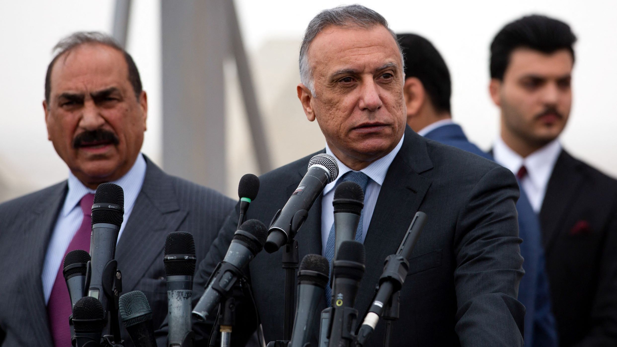 L'image montre un groupe d'hommes lors d'une conférence de presse. Au premier plan, deux hommes sont debout devant plusieurs microphones. L'un d'eux, qui se tient à gauche, porte un costume rayé et semble s'adresser à l'audience. L'autre homme, au centre, est également en costume et adopte une pose sérieuse, regardant au loin. En arrière-plan, d'autres hommes en costumes sont visibles, ajoutant à l'ambiance formelle de l'événement. L'environnement semble légèrement brumeux.