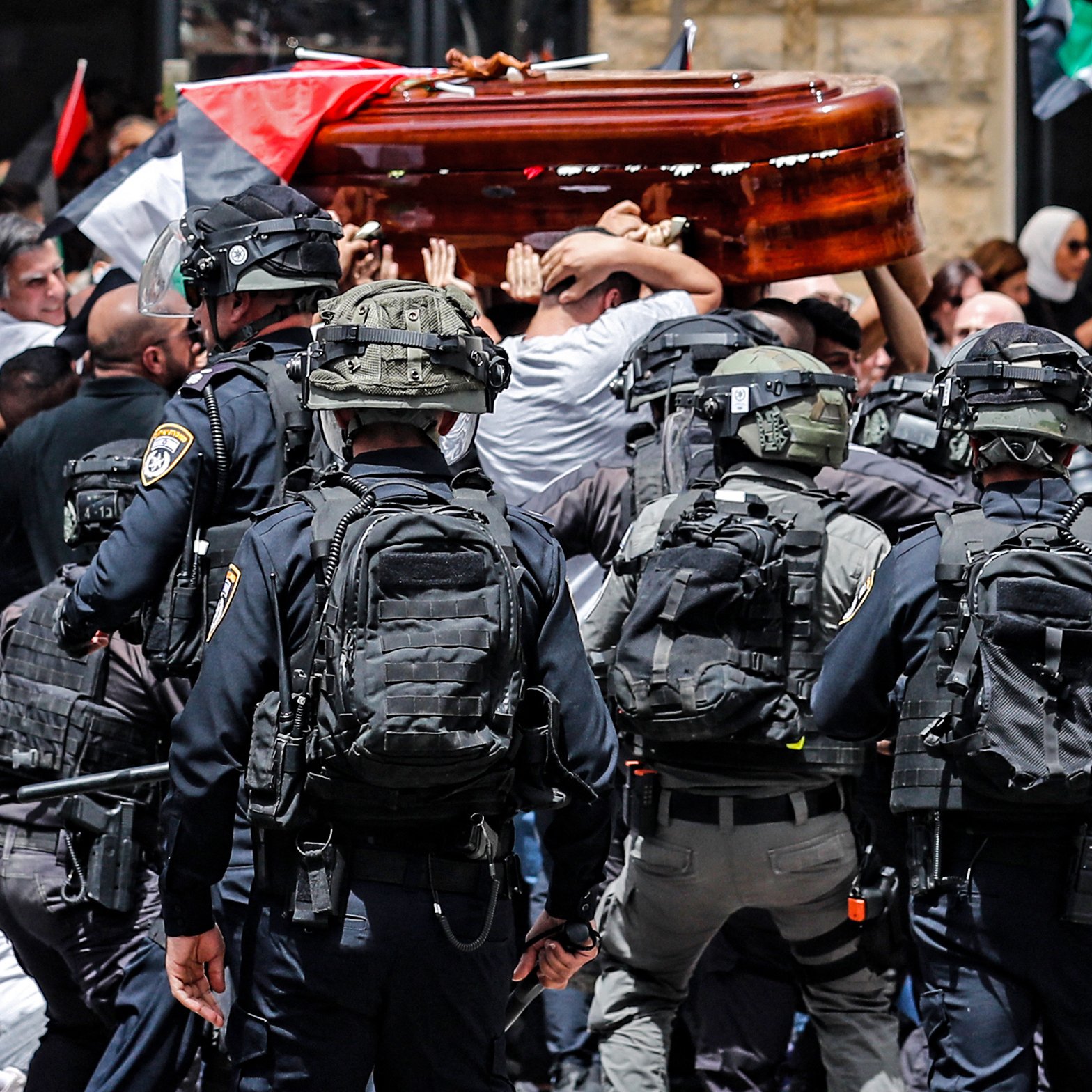 L'image montre une scène de tension où un groupe de personnes porte un cercueil en bois, tandis qu'une foule aux drapeaux palestiniens est présente. Des policiers en uniforme et équipés apparaissent en première ligne, semblant gérer la situation. L'atmosphère semble chargée d'émotion, et la scène est marquée par le contraste entre le deuil et les forces de l'ordre.