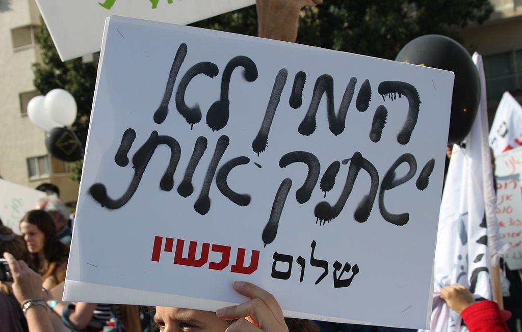 L'image montre une personne tenant une pancarte pendant une manifestation. La pancarte est écrite en hébreu, et on peut voir des slogans ou des messages gravés dessus. En arrière-plan, il y a d'autres manifestants et des ballons, ce qui suggère un rassemblement public. L'ambiance semble être celle d'une protestation, avec une atmosphère engagée.