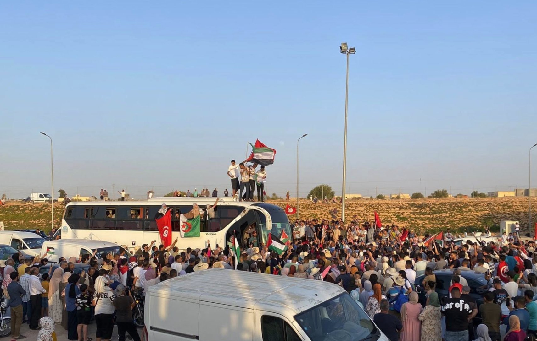 A large crowd gathers, waving flags and celebrating, with vehicles parked around.