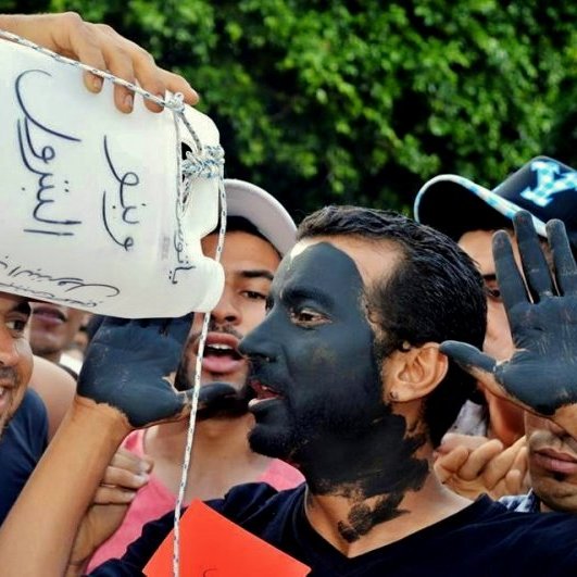L'image montre un groupe de personnes en train de participer à une manifestation ou un rassemblement. Au premier plan, deux hommes attirent l'attention. L'un d'eux porte un récipient en plastique sur la tête, tandis que l'autre a le visage peint en noir. Ils semblent discuter ou se divertir, entourés de plusieurs autres personnes qui participent à l'événement. L'ambiance paraît animée et engagée, mettant en avant des expressions de solidarité ou de protestation.