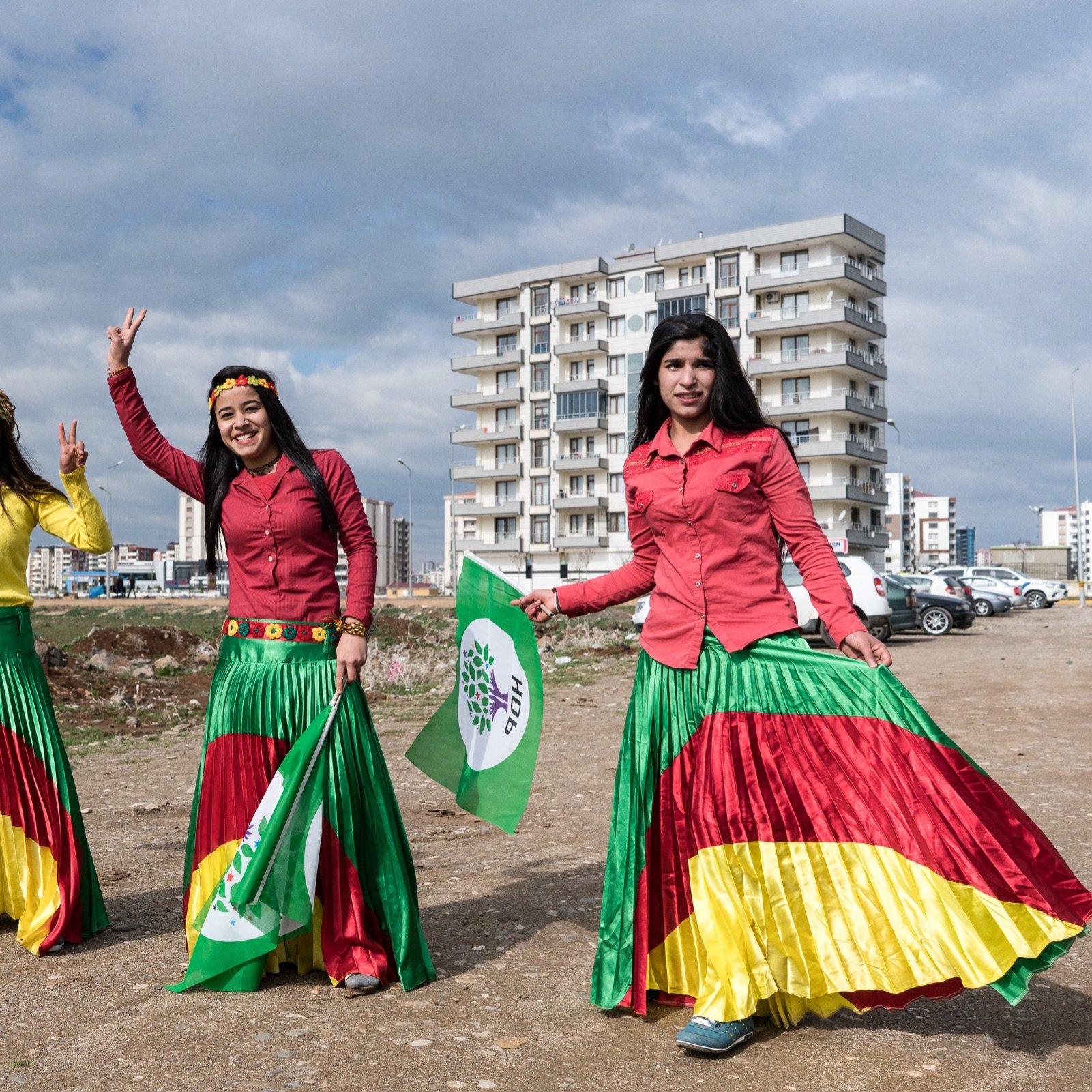 L'image montre trois femmes portant des jupes colorées, affichant des drapeaux et symboles. Elles sont entourées d'un paysage urbain, avec des bâtiments en arrière-plan et un terrain dégagé. Les femmes semblent célébrer un événement, affichant des sourires et des gestes de victoire. Les couleurs vives de leurs vêtements et des drapeaux évoquent un sentiment de fierté culturelle.
