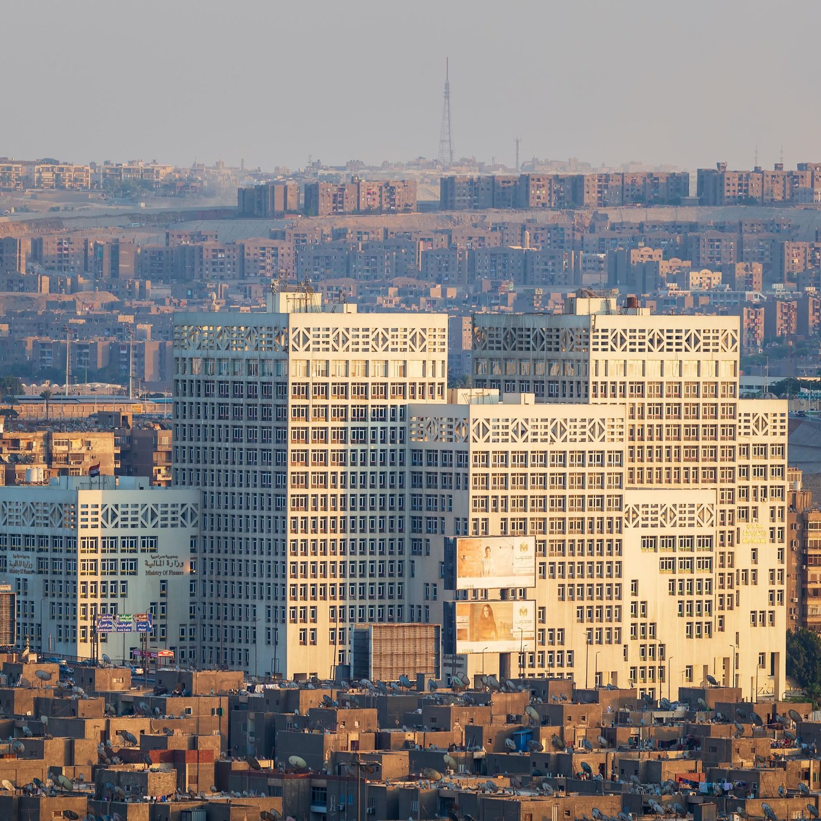 L'image montre un paysage urbain avec plusieurs bâtiments modernes au premier plan, qui se distinguent par leur architecture unique et leurs façades vitrées. En arrière-plan, on aperçoit une vaste étendue de constructions résidentielles plus anciennes, avec des habitations compactes et souvent uniformes. La lumière du soleil semble éclairer les bâtiments modernes, créant un contraste avec le reste de la ville, particulièrement à l'heure dorée du matin ou du soir. Le ciel apparaît clair, ce qui met en valeur la scène urbaine.