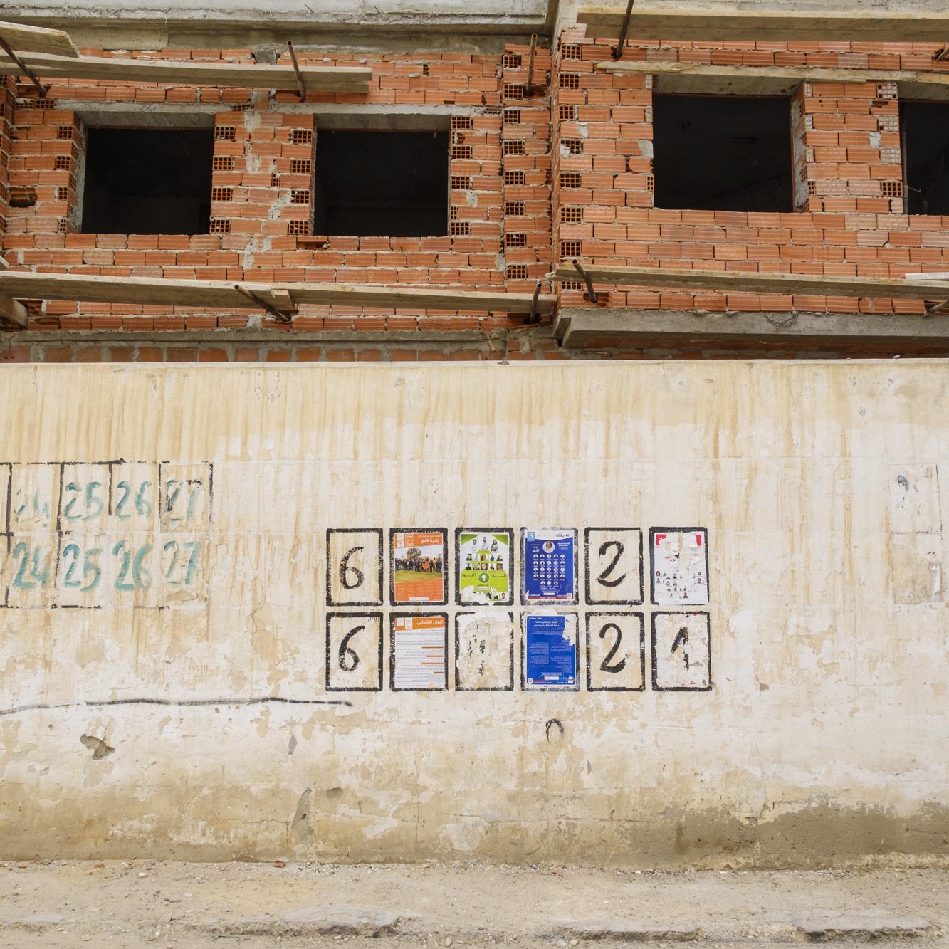 L'image montre un mur en béton, partiellement recouvert de peinture, qui semble faire partie d'un chantier de construction. Sur ce mur, il y a plusieurs cases carrées, probablement marquées pour indiquer des numéros ou des étapes de progression. À la centre, quelques affiches et documents sont accrochés, fournissant des informations peut-être liées au chantier. En arrière-plan, on aperçoit un bâtiment en construction avec des briques exposées et des fenêtres incomplètes, ce qui suggère un contexte de travaux en cours.