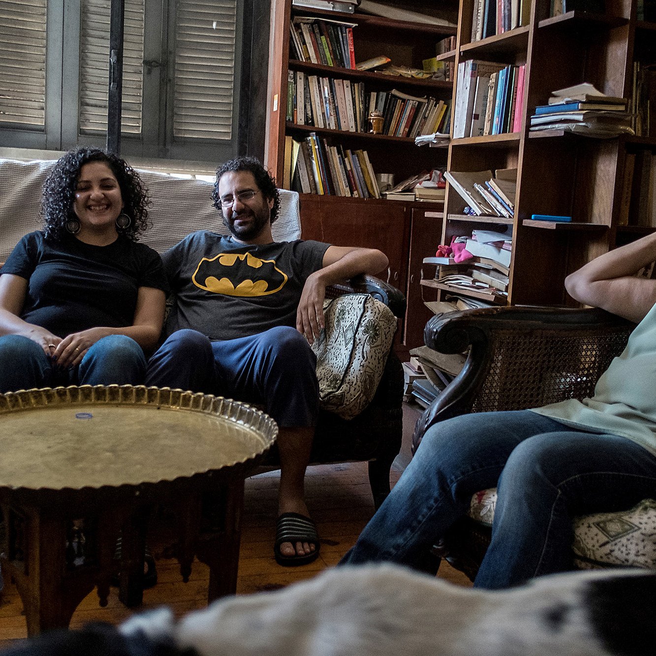 L'immagine mostra un ambiente domestico accogliente, con un soggiorno arredato in modo informale. Ci sono tre persone sedute su poltrone e divani. Due di loro sorridono, mentre un'altra donna, con capelli grigi e ricci, appare rilassata. In mezzo c'è un tavolino rotondo di metallo. Sullo sfondo, ci sono librerie piene di libri, che conferiscono un'atmosfera calda e familiare all'ambiente. Sul pavimento, si intravede un cane.