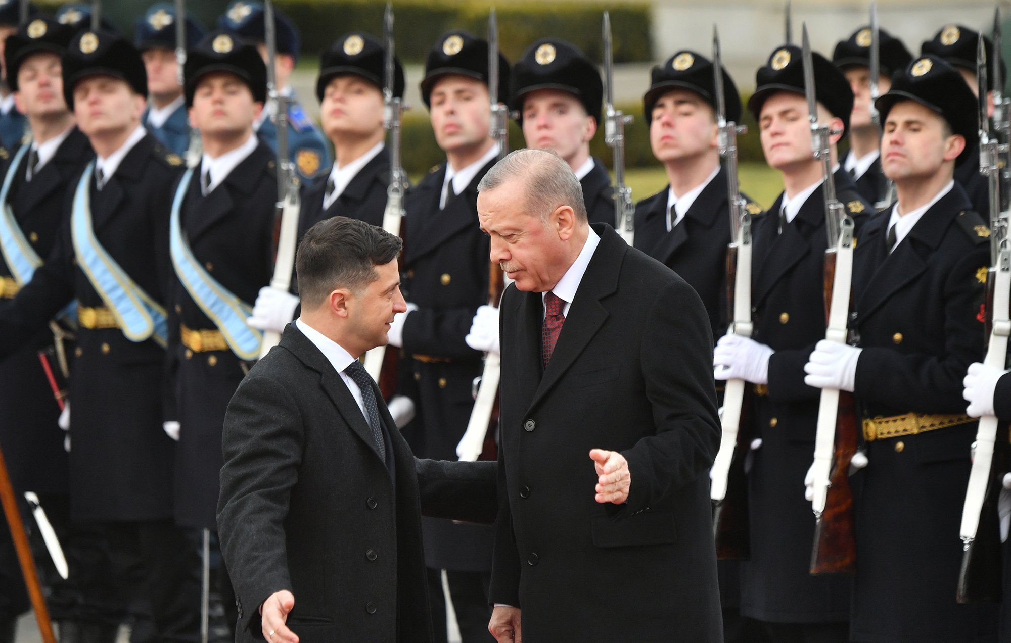 تظهر الصورة مجموعة من الجنود في الزي الرسمي يتواجدون في تشكيل عسكري أمام شخصيتين بارزتين. الشخصيات تبدو وكأنها تتحدثان معًا، حيث يبدو أحدهما في حالة نقاش مع الآخر. الجنود خلفهم يقفون بانضباط، بينما يتم عرض الأسلحة بشكل منظم. الأجواء تعكس طابعًا رسميًا وجديًا للحدث.