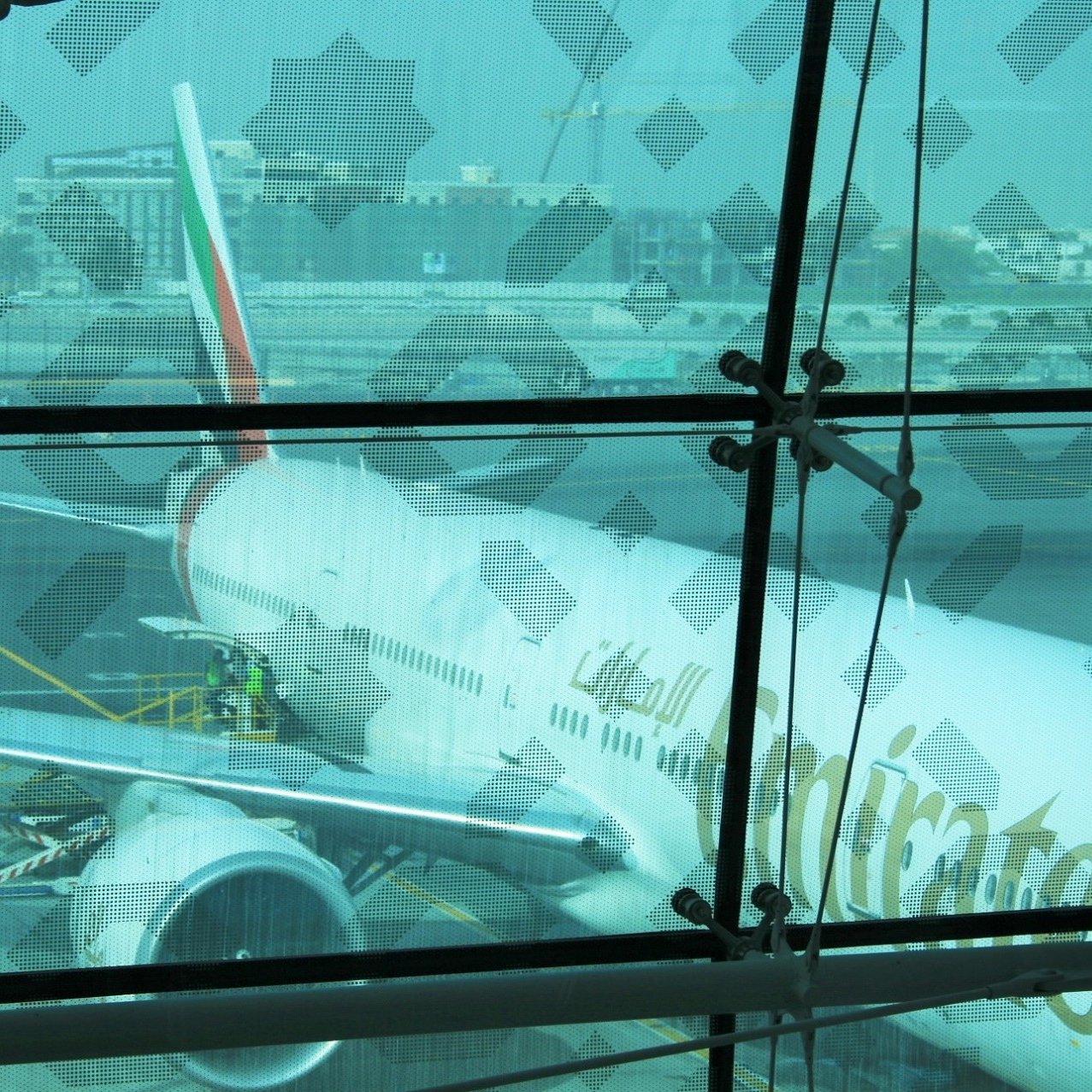 The image shows an airplane belonging to Emirates Airlines parked at an airport. The aircraft appears to be a large model, likely a Boeing 777, as indicated by the shape of the fuselage and engines. You can see maintenance personnel working on the plane, and the background features airport buildings and infrastructure. The scene is framed by a glass wall, which has a patterned overlay, adding an artistic element to the view. The overall atmosphere seems busy and operational, typical for a major airport.