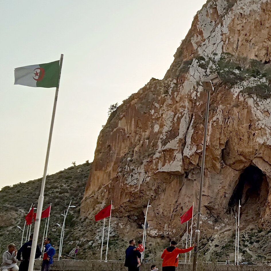 L'image montre une imposante falaise avec une grande ouverture, probablement une grotte, qui se trouve à sa base. Des drapeaux algériens flottent autour, ajoutant une touche de couleur au paysage. Au premier plan, on peut voir des personnes marchant près des drapeaux. Le ciel est teinté de lumière douce, suggérant peut-être un lever ou un coucher de soleil. L'ensemble évoque un lieu naturel remarquable avec une signification culturelle ou historique.