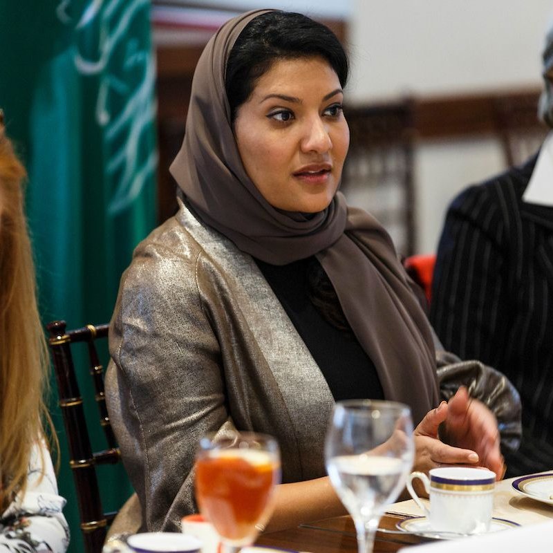 L'image montre un groupe de femmes assises autour d'une table, en train de discuter. Une femme au centre porte un hijab et un vêtement aux couleurs neutres, semblant engagée dans la conversation. À sa gauche, une autre femme aux cheveux longs et blonds, légèrement penchée, écoute attentivement. À droite, une femme en tailleur noir et blanc observe également. Sur la table, on peut voir des verres de boisson et de la vaisselle, avec des éléments de nourriture. L'environnement semble formel, propice à une réunion ou un déjeuner d'affaires.