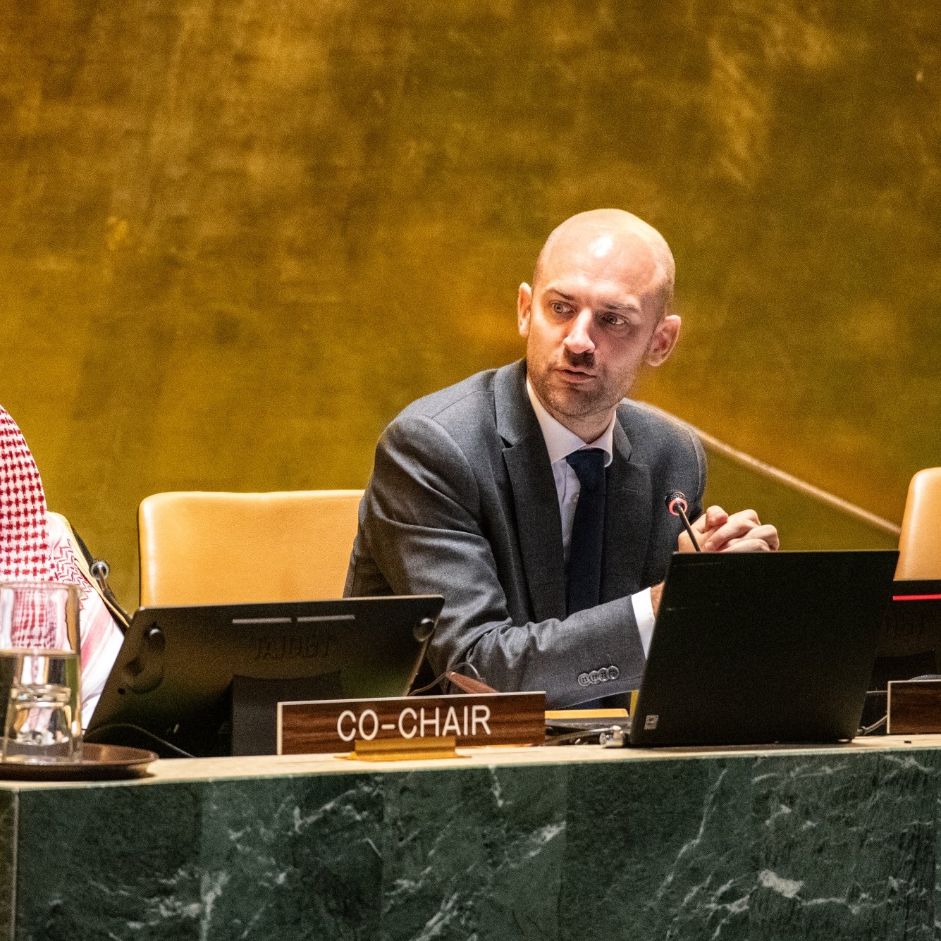 Two men at a conference table, one in traditional attire, discussing on a stage with microphones.