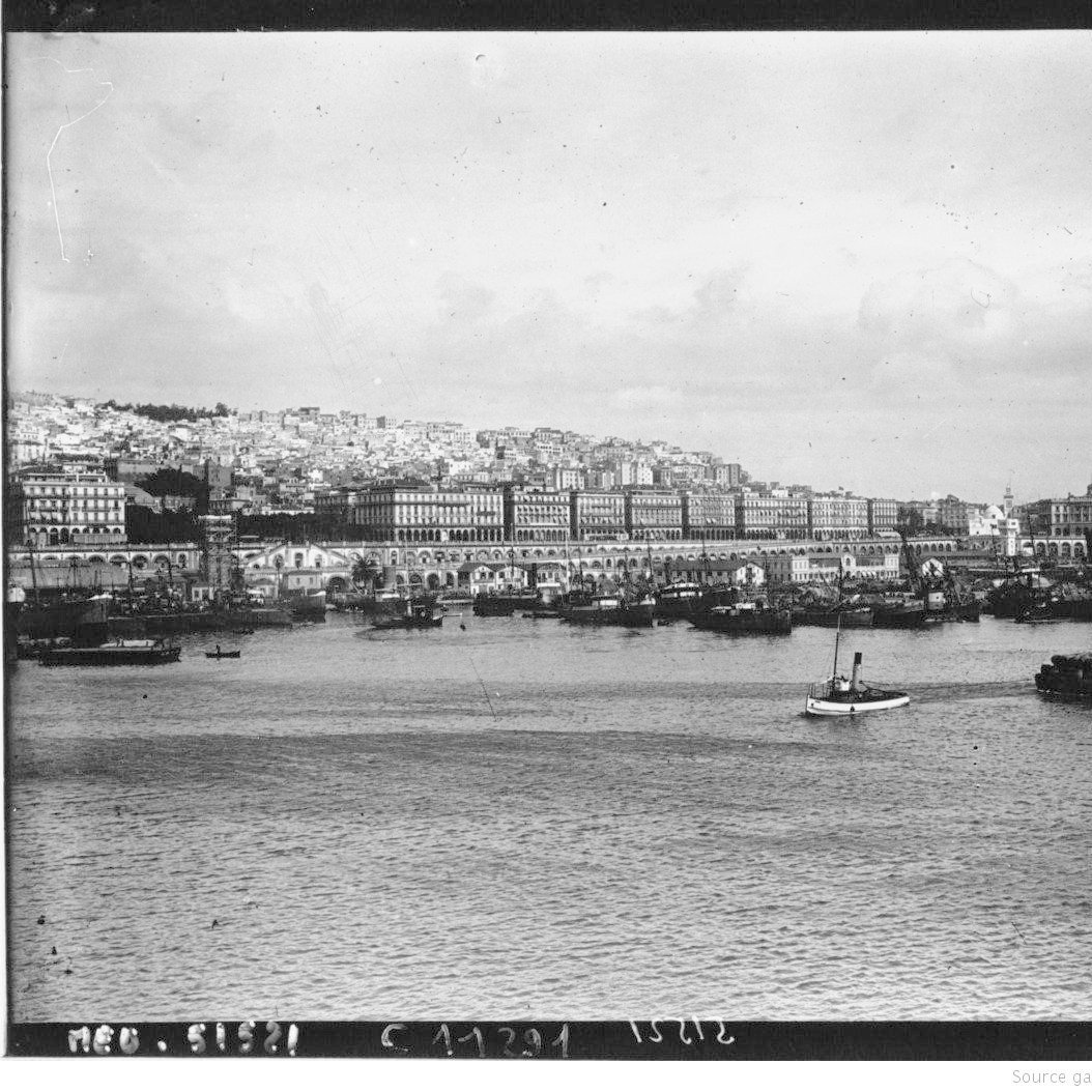 L'image représente un port où l'on peut voir plusieurs bateaux amarrés. Au fond, une ville s'étend, avec des bâtiments alignés sur la côte. Le ciel est nuageux, et l'eau calme reflète la scène. Les collines derrière la ville sont habitées, ajoutant à la densité urbaine de l'endroit. L'ambiance générale semble nostalgique, probablement en raison du style photographique ancien.