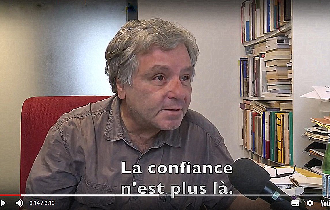 L'image montre un homme assis devant un bureau, dans un bureau plutôt ordonné avec des livres en arrière-plan. Il parle dans un microphone et semble discuter d'un sujet important. Sur l'écran, on peut voir le sous-titre qui indique : "La confiance n'est plus là." L'éclairage est neutre, et l'homme a une expression sérieuse.