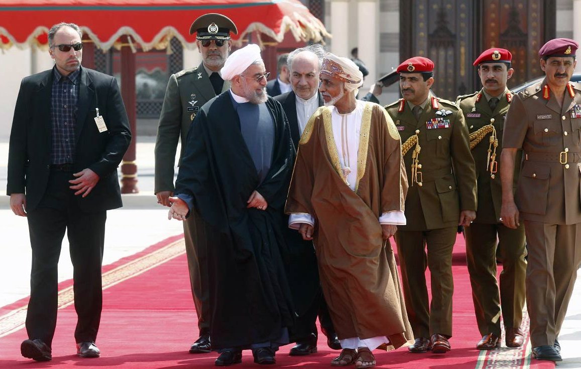 L'image montre un groupe de personnes marchant sur un tapis rouge. Au centre, deux hommes en discussion, l'un portant une robe noire et l'autre une robe traditionnelle d'Oman. Ils sont entourés de plusieurs membres de la sécurité et d'officiers en uniforme. Le cadre est formel, avec un décor architectural en arrière-plan qui suggère un événement diplomatique ou officiel.