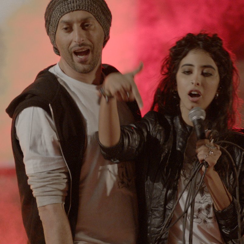 L'image montre des artistes en pleine performance musicale, avec une ambiance vibrante et colorée. Au centre, une femme chante dans un microphone, accompagnée d'un homme à ses côtés qui semble participer activement à la performance. En arrière-plan, d'autres musiciens se joignent à l'animation, entourés de lumière et de fumée, créant une atmosphère dynamique et festive. Les expressions des artistes indiquent de l'enthousiasme et de l'énergie.