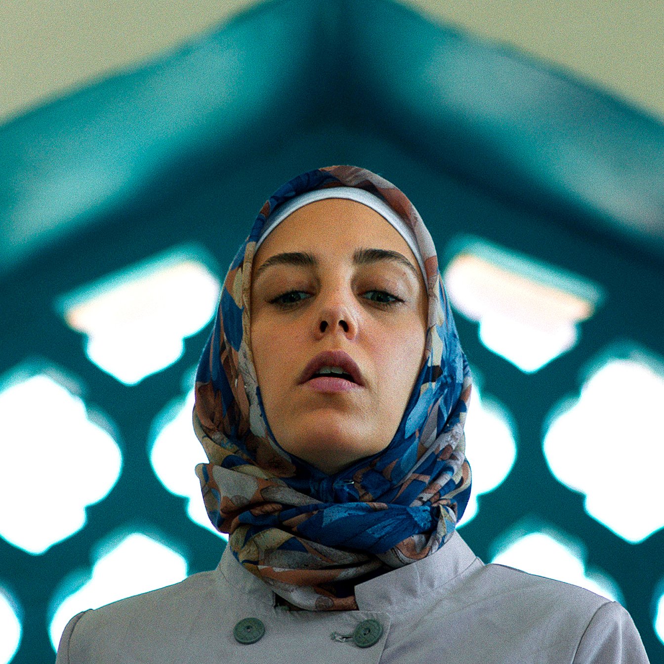 L'image montre une femme portant un hijab coloré, se tenant dans un cadre architectural. La lumière met en valeur son visage et le fond présente des motifs géométriques typiques des designs islamiques, avec des arches et des vitraux. Son expression est sérieuse, ce qui ajoute à la profondeur de la scène.