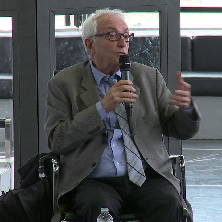 Il s'agit d'un homme assis sur une chaise, tenant un microphone, probablement en train de parler ou de donner une présentation. Il porte une veste et une cravate, et l'environnement semble être un espace public ou une salle de conférence. Des éléments modernes de design sont visibles en arrière-plan.