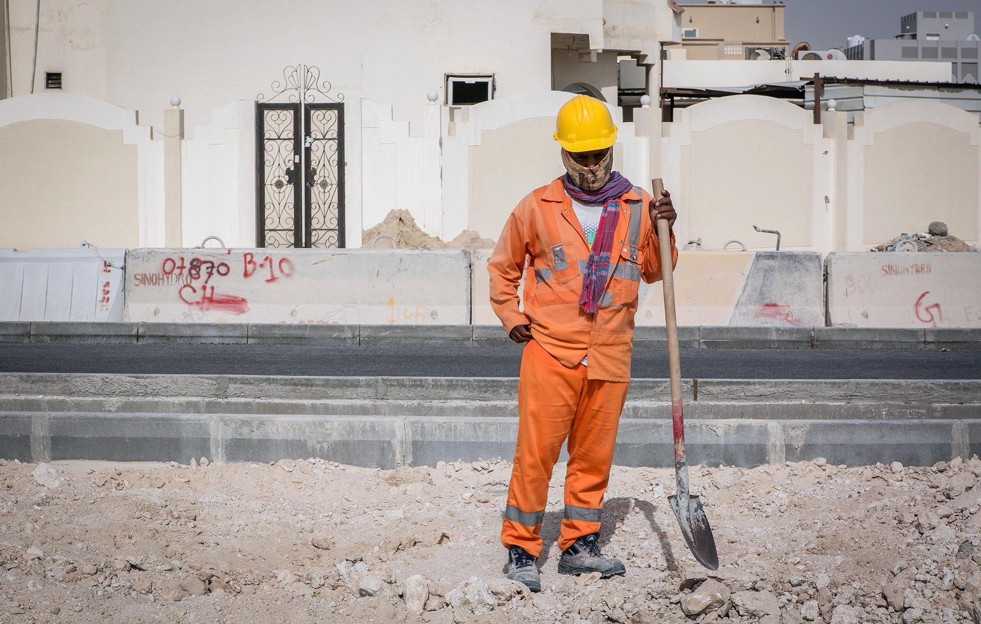 L'image montre un homme qui travaille sur un chantier de construction. Il porte un uniforme orange, ainsi qu'un casque de sécurité jaune. Il semble tenir une pelle, regardant vers le sol avec une posture réfléchie. En arrière-plan, on peut voir des murs de construction et des graffitis sur des surfaces environnantes. Le site semble en cours de développement, avec des matériaux de construction dispersés au sol.
