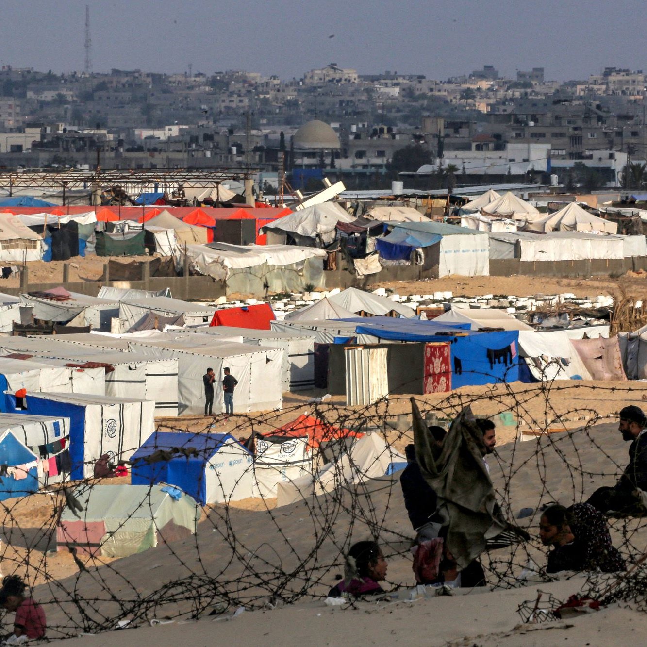 L'immagine rappresenta un campo profughi, con numerosi tendoni di vari colori, come blu, bianco e arancione, disposti in un'area sabbiosa. Sullo sfondo si intravedono edifici e una città, mentre una rete di recinzione sembra delimitare il campo. In primo piano, si possono vedere alcune persone sedute, creando un senso di comunità nonostante le difficili condizioni di vita. La scena evoca una forte emozione, evidenziando la realtà delle persone costrette a vivere in tale situazione.