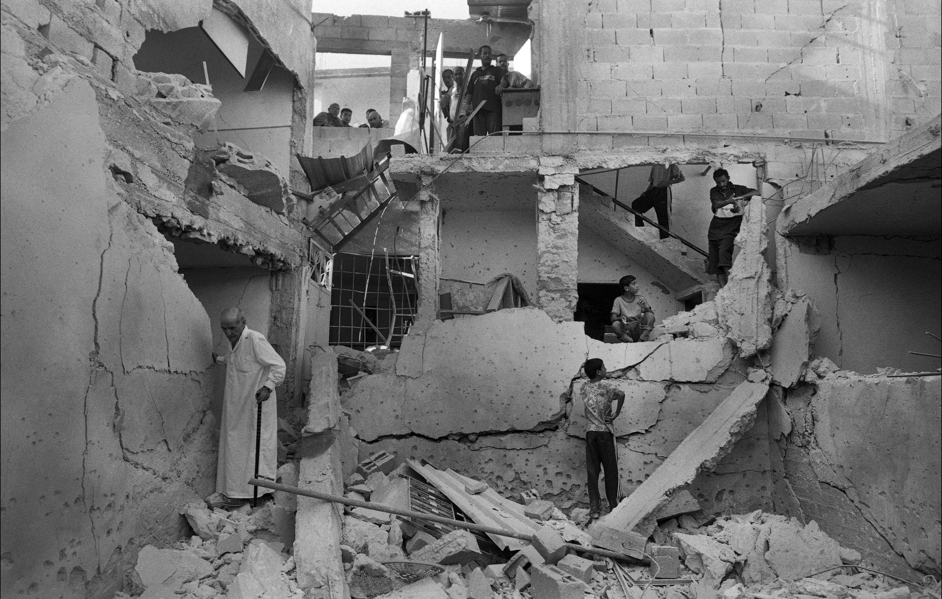 L'image montre un bâtiment en ruines, visiblement endommagé par un conflit ou un désastre. Les murs sont effondrés et des débris jonchent le sol. Plusieurs personnes se trouvent dans l'espace, certains semblent fouiller parmi les débris, tandis que d'autres observent la scène depuis un étage supérieur. L'ambiance est sombre et témoigne des conséquences dévastatrices sur la vie humaine et les infrastructures.