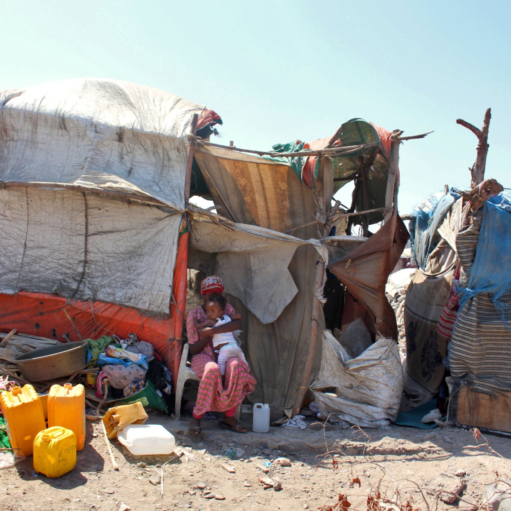 L'image montre une habitation informelle construite avec des matériaux comme des bâches en plastique et des morceaux de toile. À l'avant, on peut voir une femme assise dans l'ombre de la structure, entourée de divers objets et contenants, probablement utilisés pour la vie quotidienne. L'environnement semble un peu désordonné, avec des débris et des matériaux épars autour de l'habitation. Le ciel est dégagé, ce qui donne une impression de chaleur.