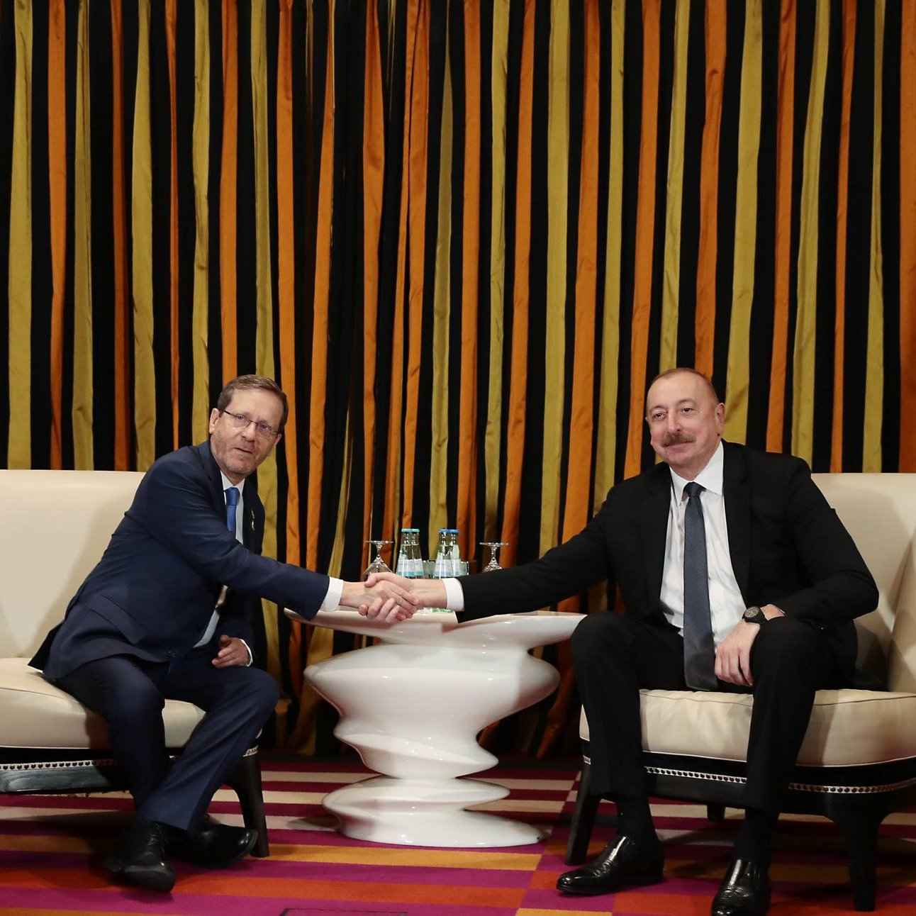 Deux hommes, en costume, se serrent la main dans un cadre élégant avec des drapeaux.