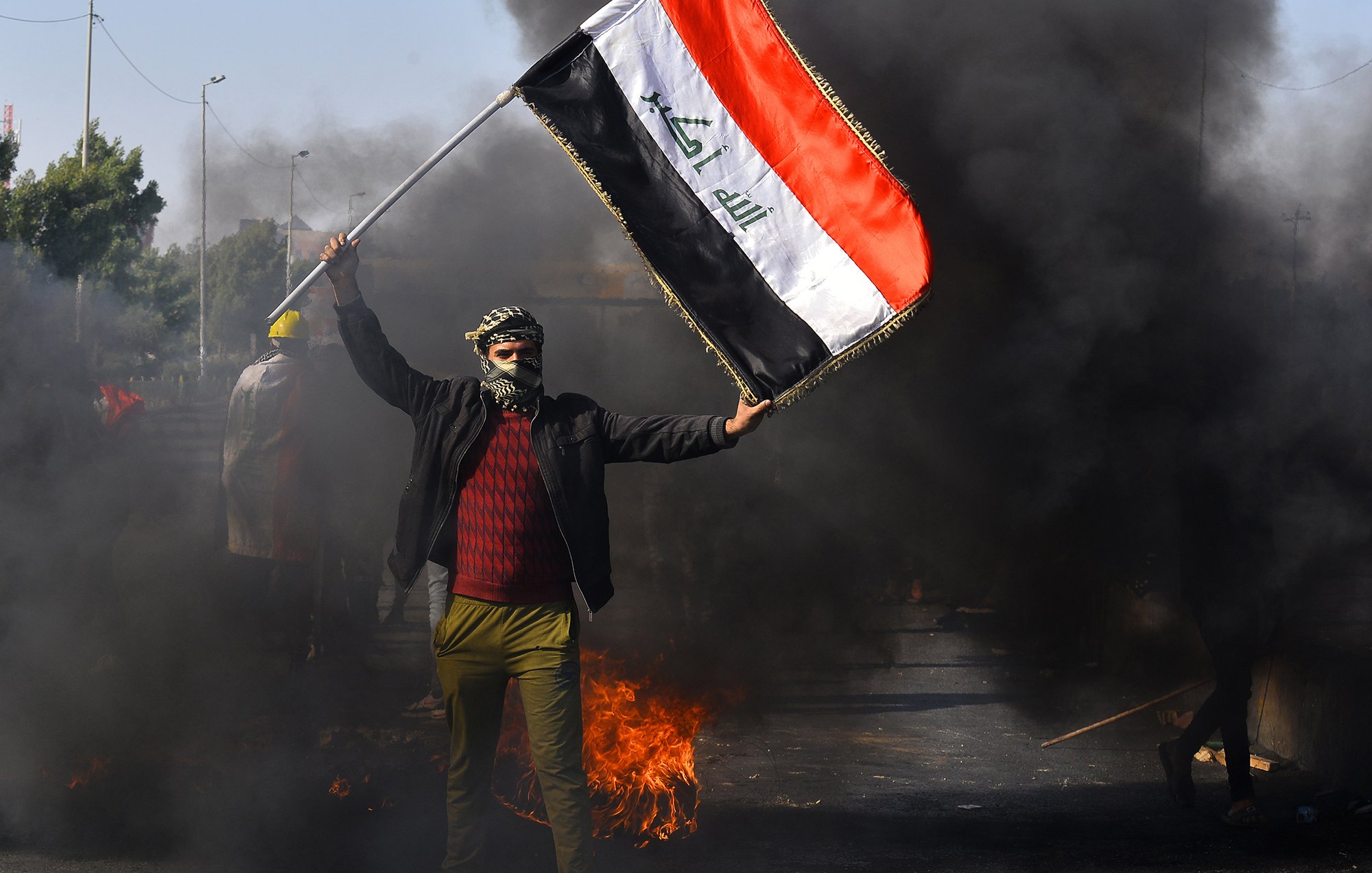 L'image montre une scène de manifestation en Irak. Un homme, portant un couvre-visage et des vêtements sombres, brandit un drapeau irakien. En arrière-plan, on peut voir des nuages de fumée noire, suggérant des feux ou des barricades sur la route. L'atmosphère semble tendue, indiquant une situation de protestation.