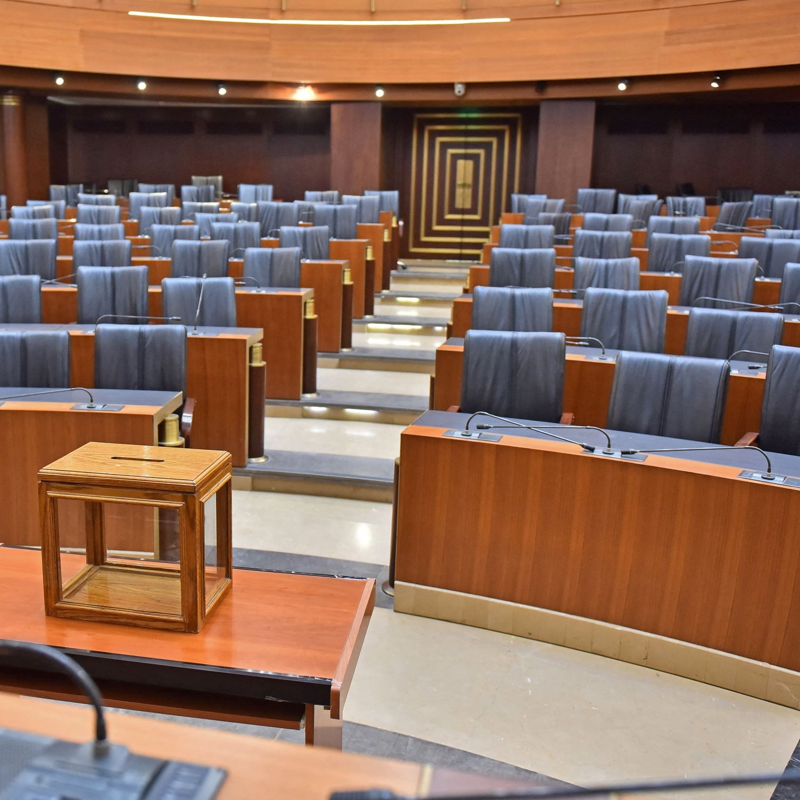 این تصویر نمایی از یک سالن جلسات یا تالار کنفرانس را نشان می‌دهد. صندلی‌های چوبی و چرمی مشکی با فاصله منظم در ردیف‌های متعدد چیده شده‌اند. در وسط تالار یک میز چوبی با یک جعبه شیشه‌ای کوچک وجود دارد. نورپردازی سالن به نظر نسبتاً ملایم است و دیوارها و سطوح چوبی به فضا حس رسمی و معتبر می‌دهند.