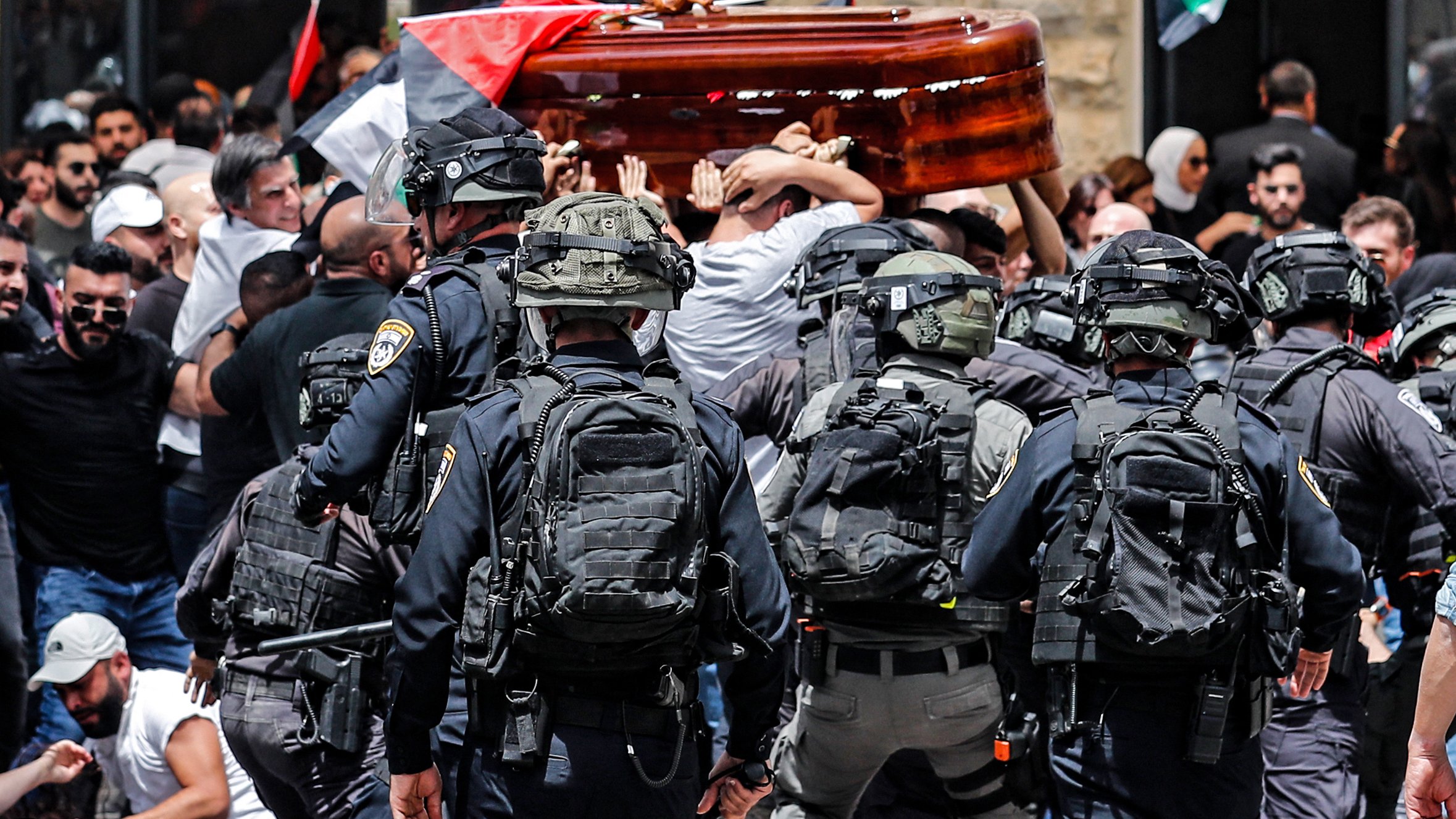 L'image montre une scène de tension où un groupe de personnes porte un cercueil en bois, tandis qu'une foule aux drapeaux palestiniens est présente. Des policiers en uniforme et équipés apparaissent en première ligne, semblant gérer la situation. L'atmosphère semble chargée d'émotion, et la scène est marquée par le contraste entre le deuil et les forces de l'ordre.