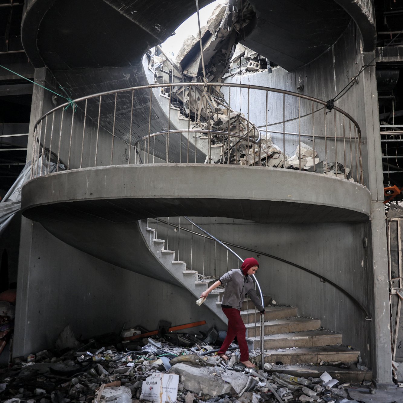 L'immagine mostra una scala a chiocciola danneggiata all'interno di un edificio in rovina. La struttura presenta macerie sparse e parte del soffitto è crollato, creando un'apertura sopra la scala. Una persona, vestita con un abbigliamento rosso e coperta da un copricapo, sta camminando sulla scala tra le macerie. L'atmosfera è quella di un luogo in crisi, con segni evidenti di distruzione e abbandono.