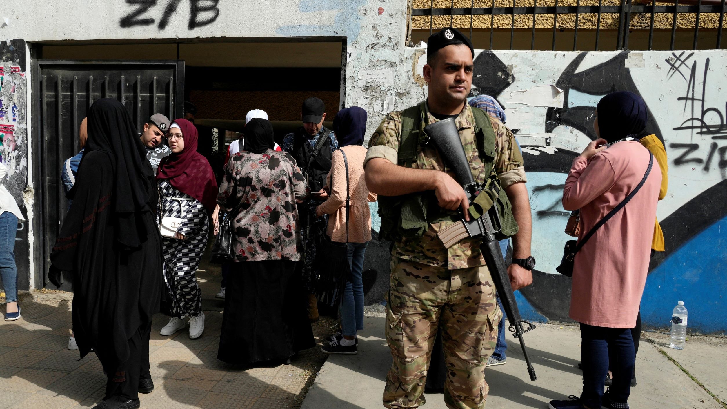 The image shows a soldier wearing military gear and holding a weapon, standing guard in front of a building. In the background, a group of people, predominantly women dressed in various styles of clothing and headscarves, are gathered near the entrance of the building. The setting appears to be an urban area, with graffiti visible on the wall behind the soldier. The atmosphere suggests a controlled environment, possibly in the context of an event or security operation.